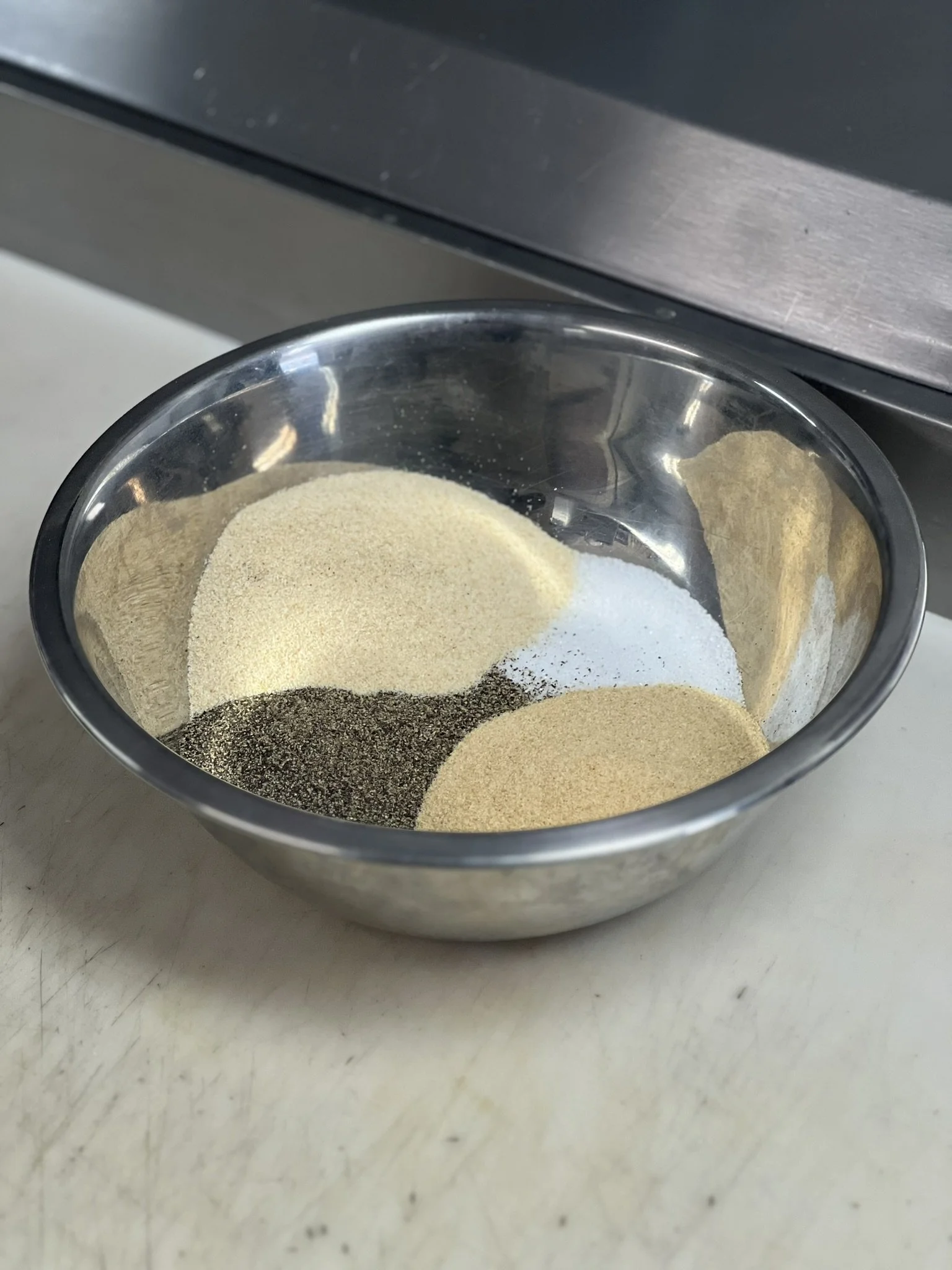 Elm Creek Beef A metal bowl containing various powdered spices, including black pepper, salt, and possibly garlic or onion powder, placed on a light-colored surface.