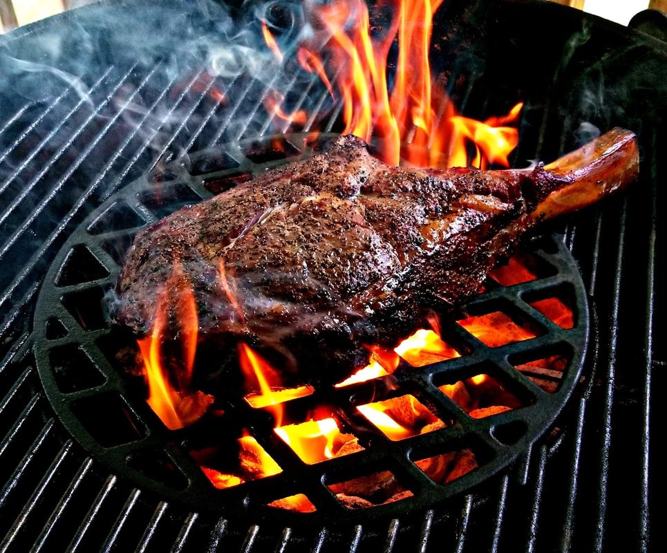 A large piece of beef steak is grilling on a barbecue, with flames and smoke around it.