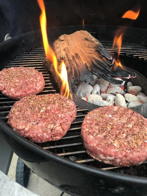 Three seasoned raw burger patties on a grill, with a piece of burning wood and hot coals in the background.