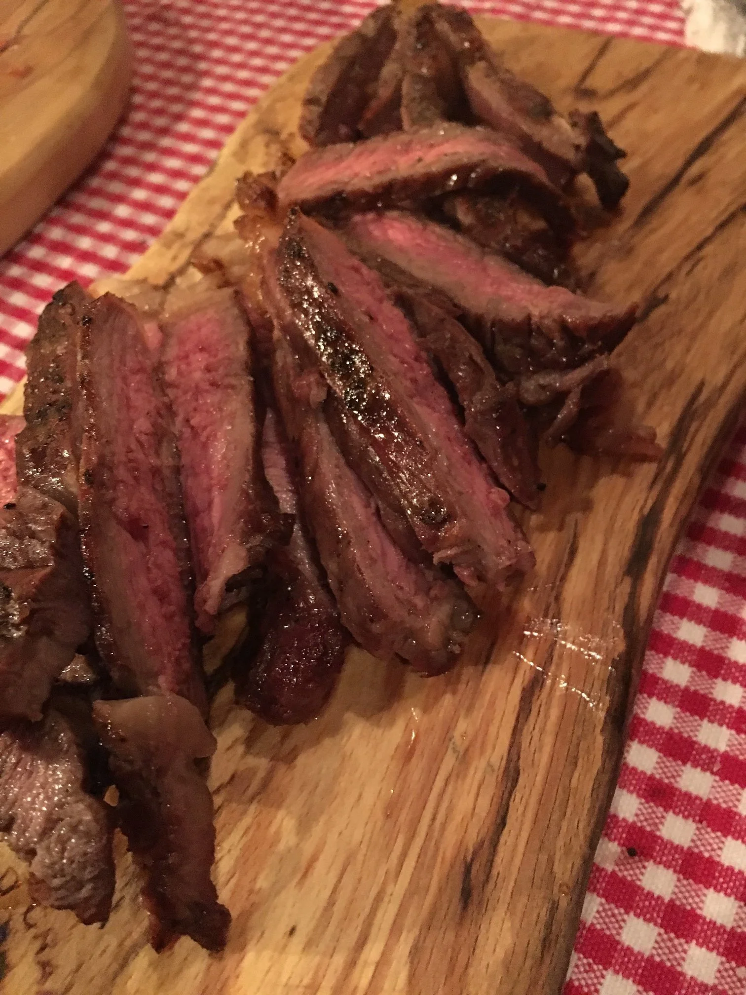 Elm Creek Beef Slices of cooked steak on a wooden cutting board with a red and white checkered tablecloth in the background.