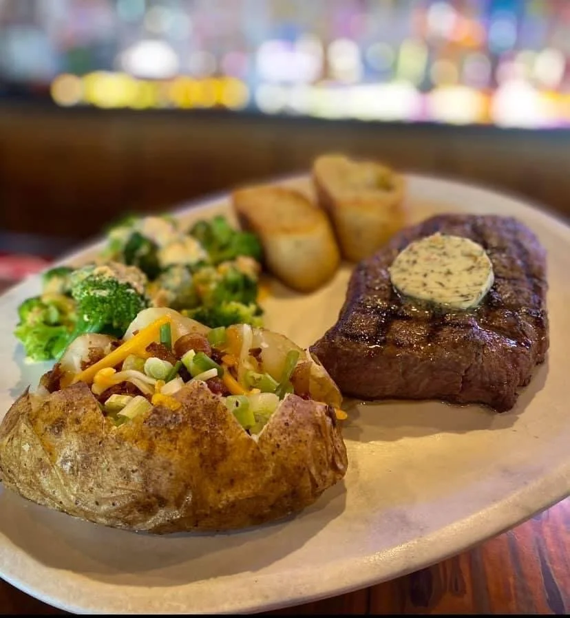 Elm Creek Beef A plate with a baked potato topped with shredded cheese and chives, a grilled steak with butter, two baked or roasted potato slices, and broccoli salad.