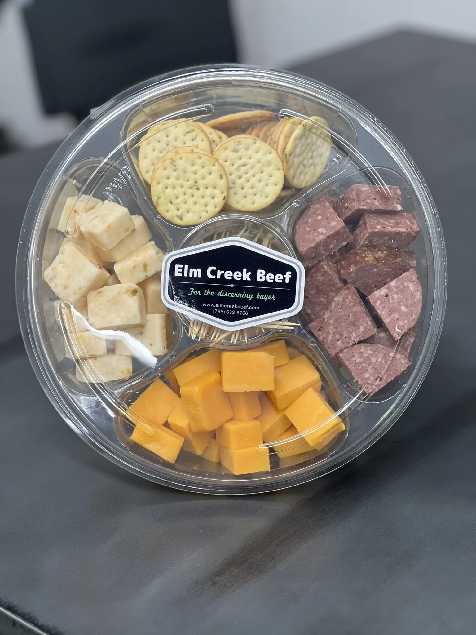 Elm Creek Beef A round plastic tray with cheese cubes, beef cuboids, crackers, and slices of cheddar cheese, labeled 'Elm Creek Beef'.