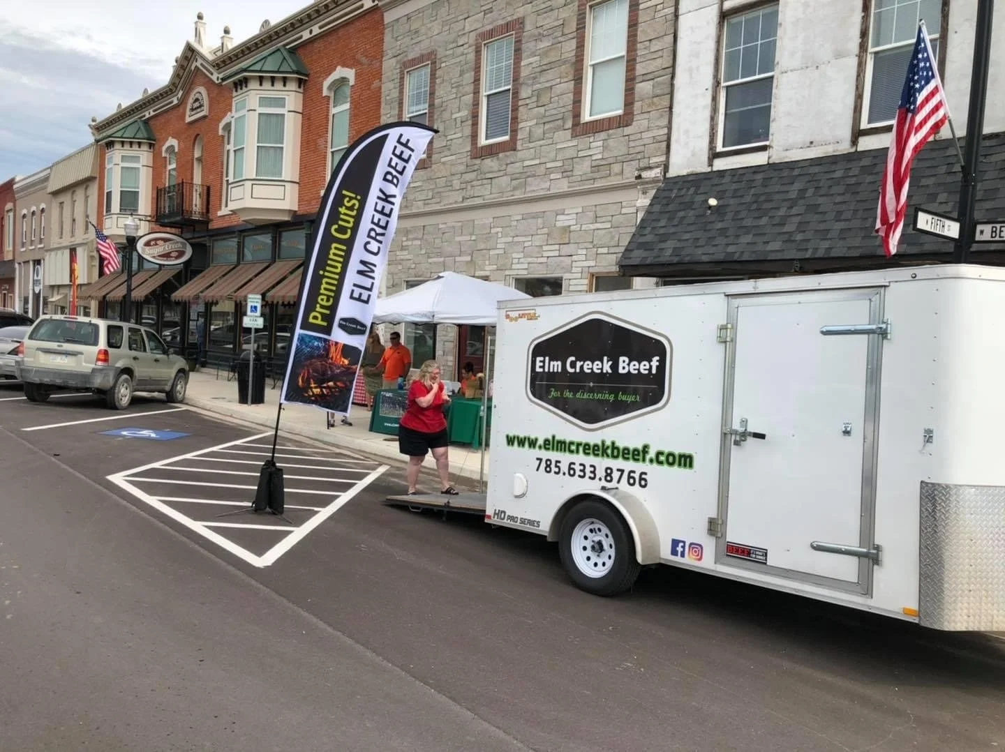 Elm Creek Beef Food truck parked on the street advertising Elm Creek Beef, with a woman in red standing nearby, and a building with storefronts and flags in the background.