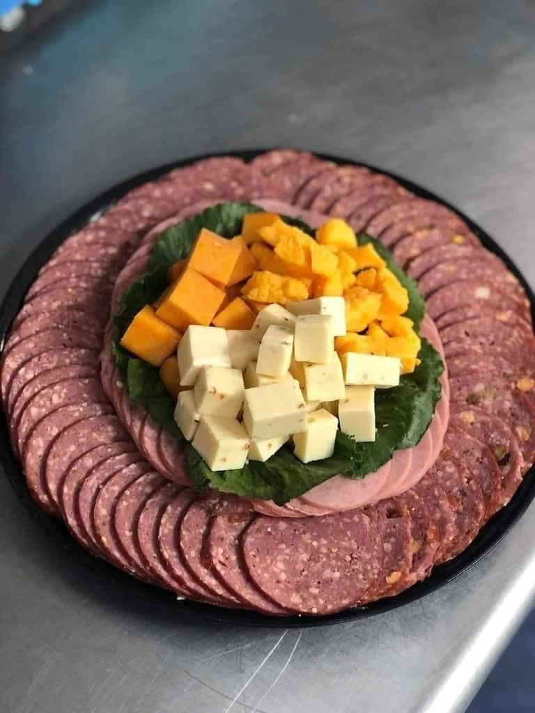 Elm Creek Beef A black platter of sliced cured salami, topped with chunks of cheddar cheese, Monterey Jack cheese, and slices of ham, arranged on a bed of green lettuce leaves.