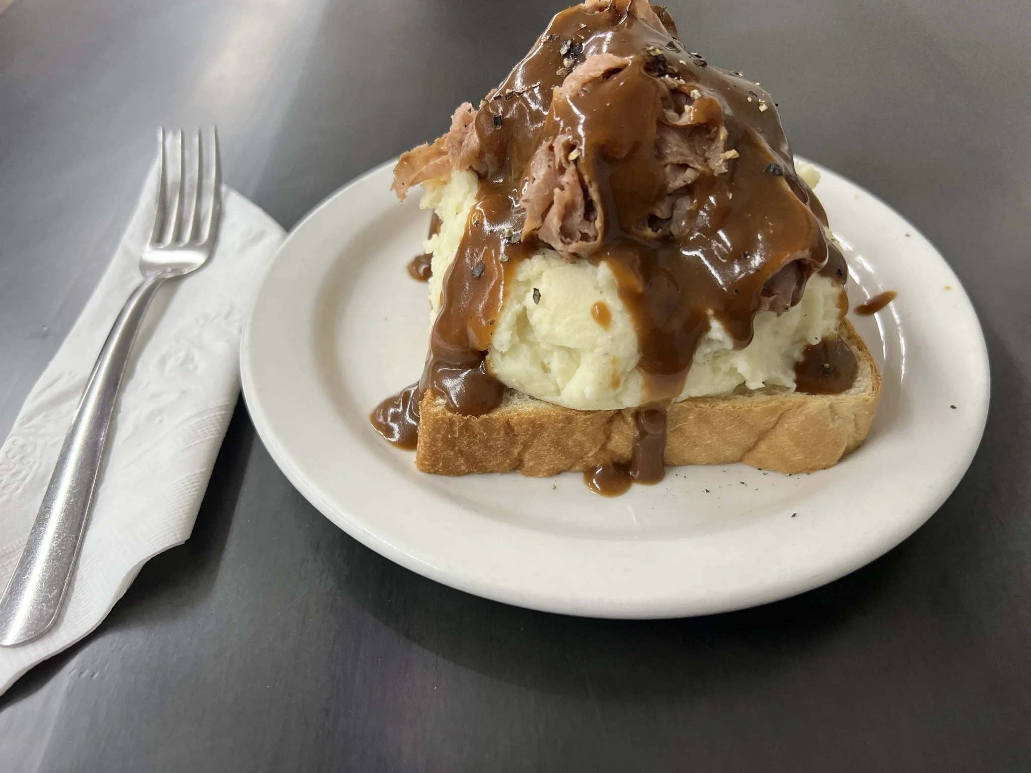 Elm Creek Beef A slice of bread topped with mashed potatoes, shredded meat, and gravy on a white plate.