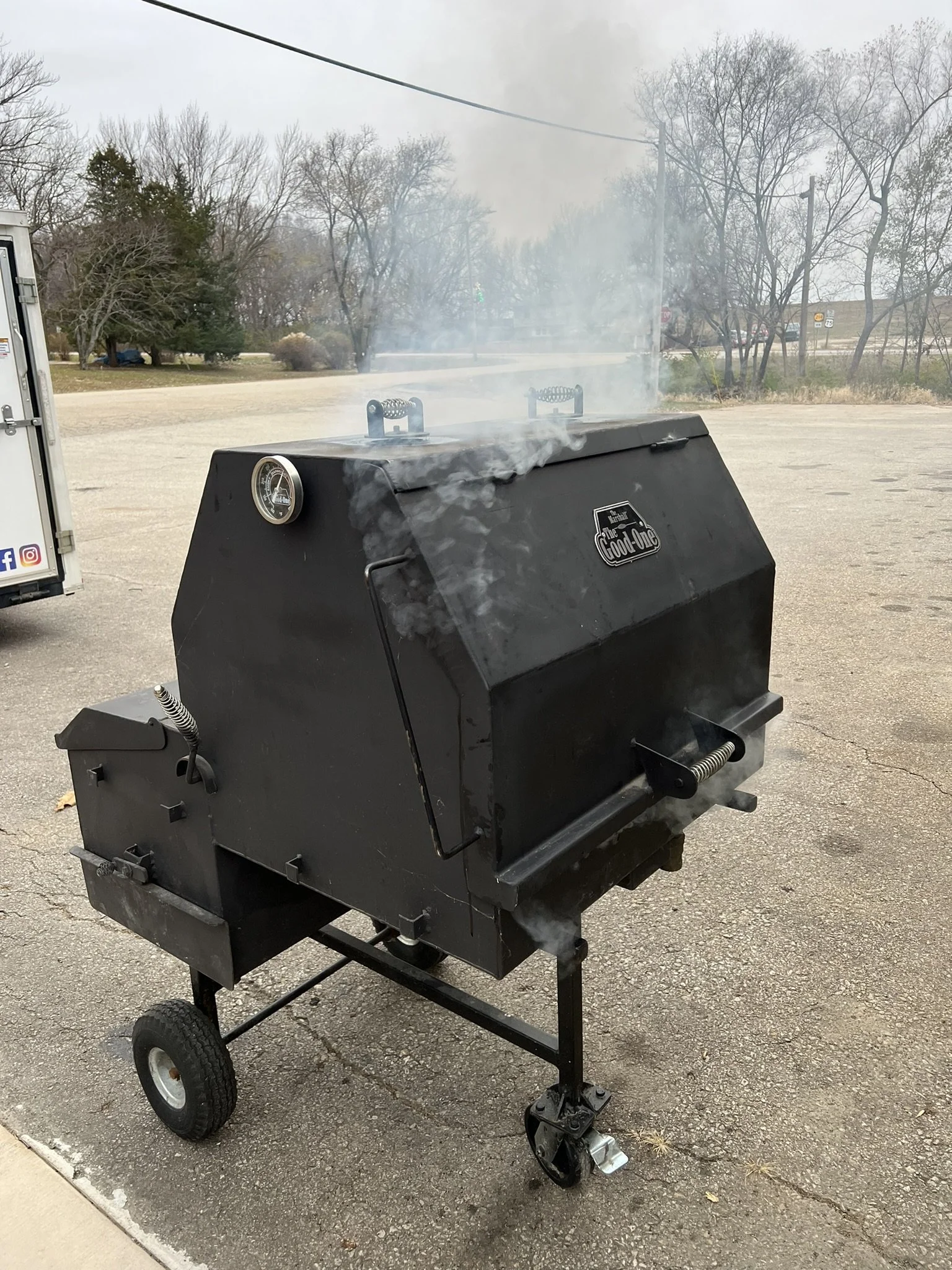 Elm Creek Beef A black smoker BBQ grill on wheels releasing smoke, located outdoors on a paved area with trees and a cloudy sky in the background.