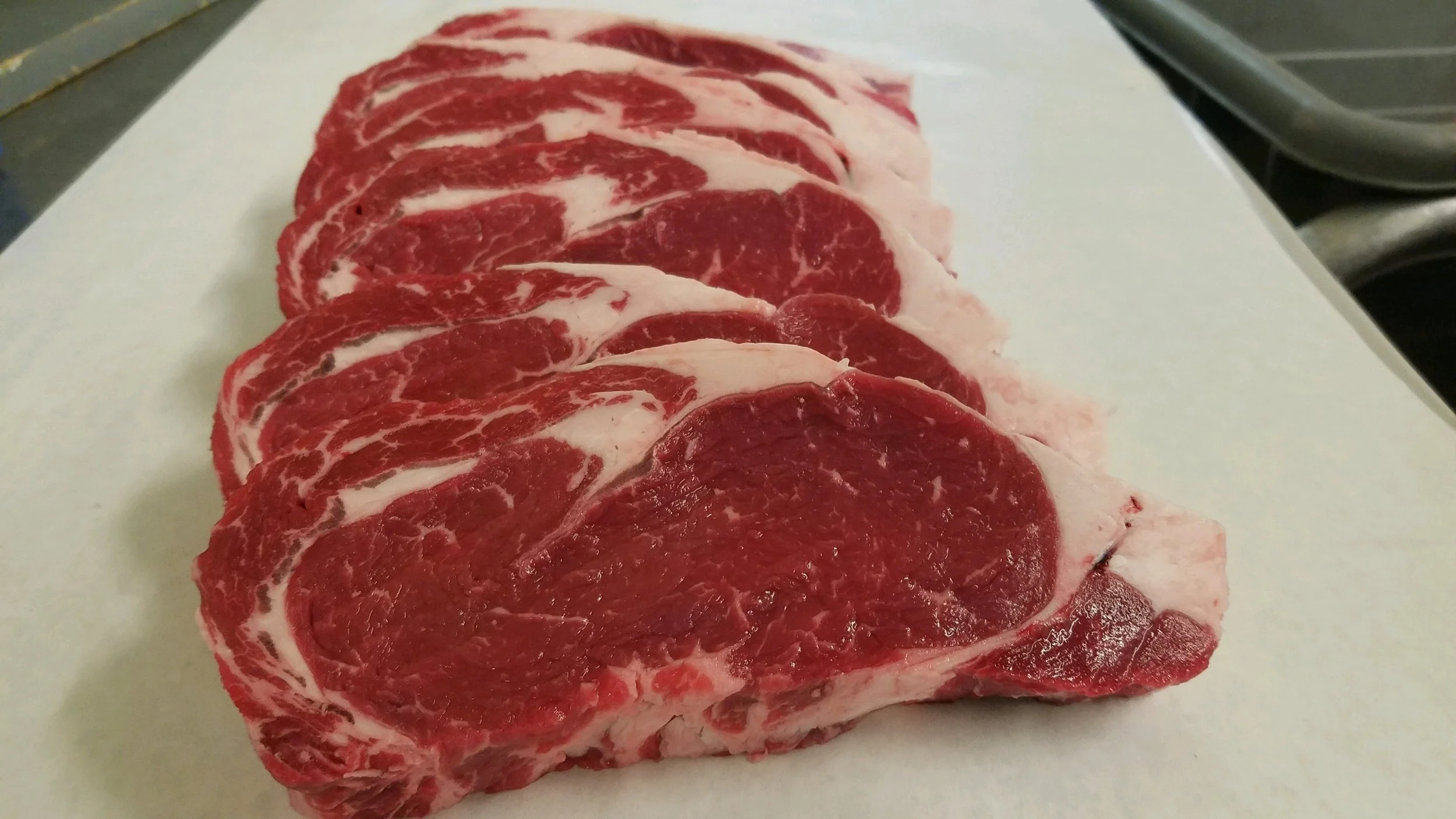 Elm Creek Beef Several raw beef steaks with marbling layer laying on white cutting board.
