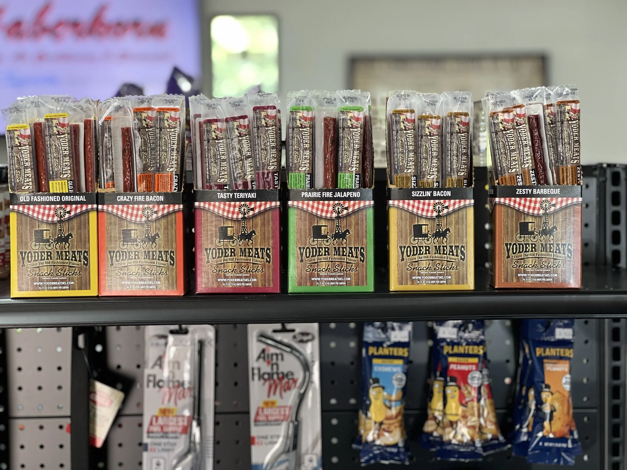 Elm Creek Beef Assorted single-serve meat snack sticks in colorful packaging on a store shelf, labeled with different flavors like Old Fashioned Original, Crazy Fire Bacon, Tasty Teriyaki, Prairie Fire Jalapeno, Sizzlin' Bacon, and Zesty Barbeque.