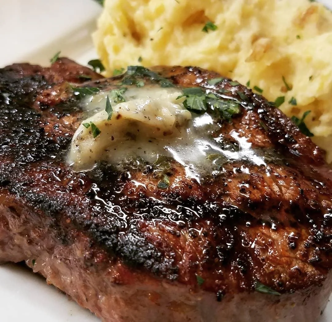 Elm Creek Beef A cooked steak topped with butter and herbs, served with a side of creamy mashed potatoes.