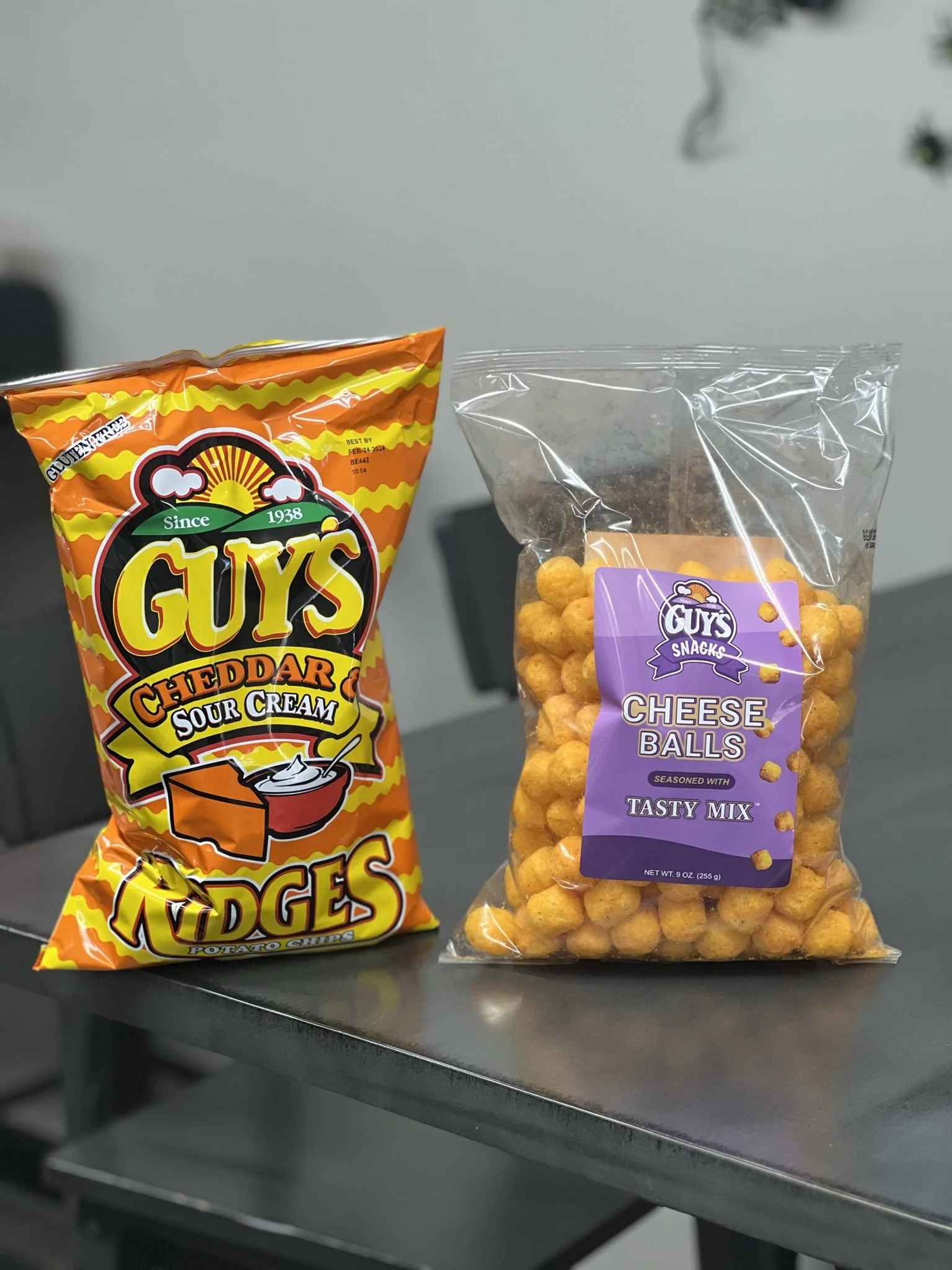 Elm Creek Beef Bag of Guy's Cheddar Sour Cream potato chips and a bag of Guy's Snack cheese balls on a dark table.