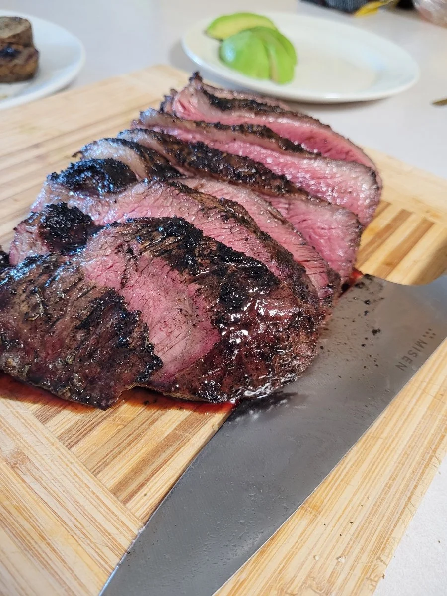 Elm Creek Beef Sliced medium-rare steak on a wooden cutting board with a carving knife, with a plate of sliced avocados in the background.
