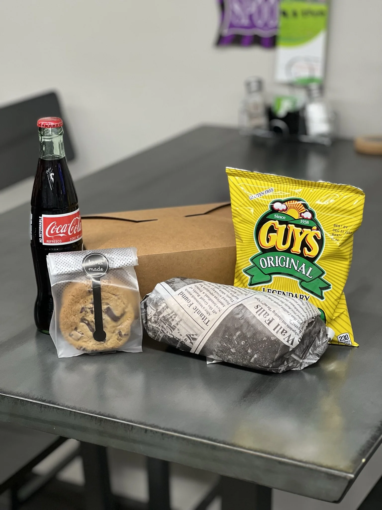 Elm Creek Beef A meal on a gray table with a Coca-Cola bottle, a cookie in a clear bag, a wrapped sandwich, a bag of Guy's Original potato chips, and a brown paper bag, with a blurred background of a black chair and some bottles.
