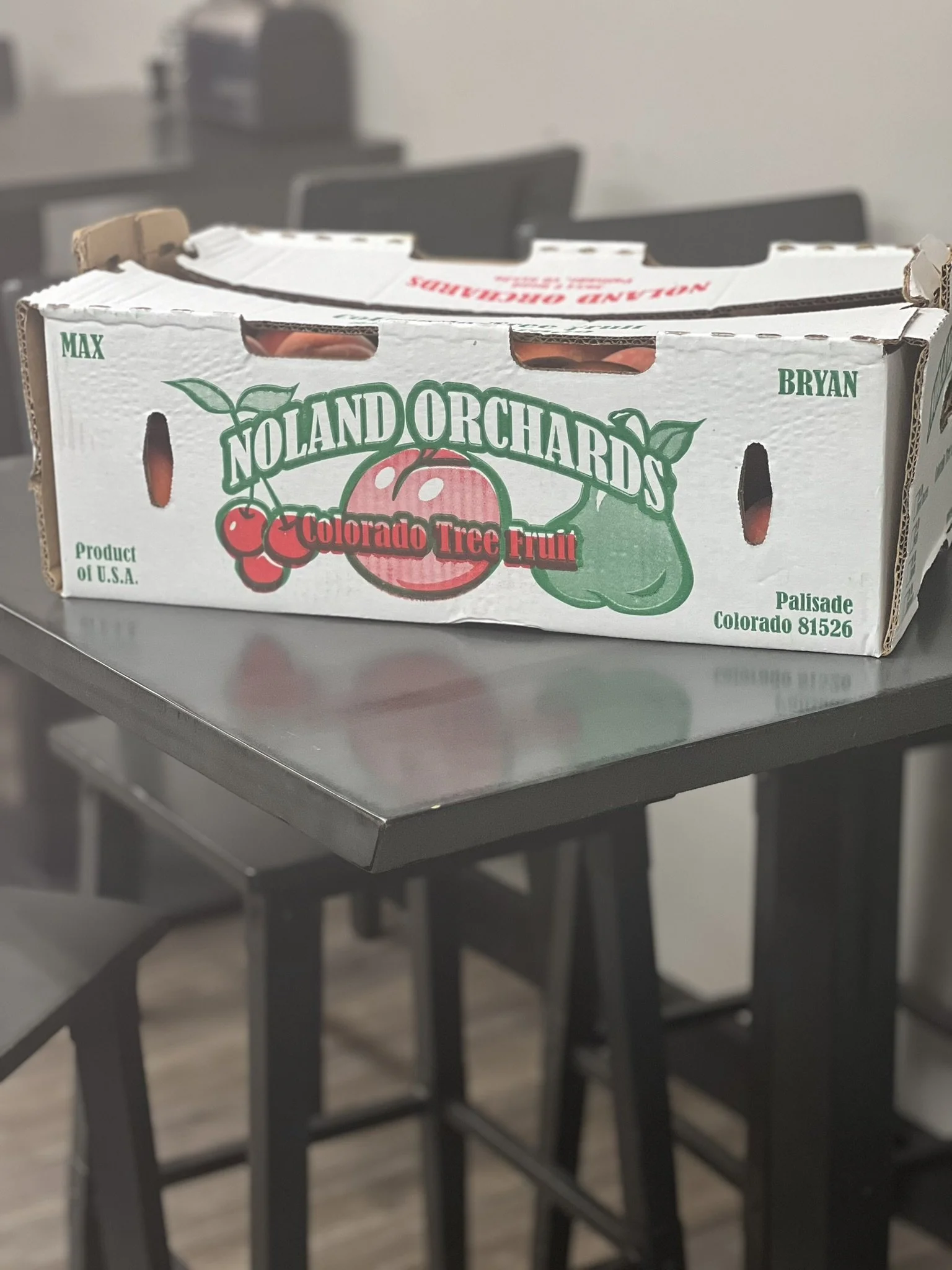 Elm Creek Beef A cardboard box of Nolan Orchards Colorado Tree Fruit on a metal table in a room with chairs and a blurred background.