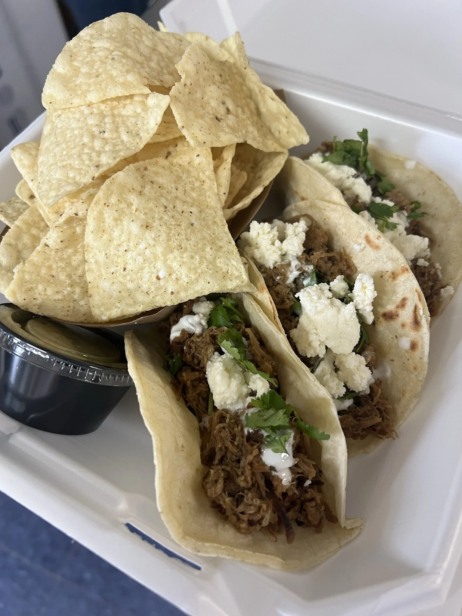 Elm Creek Beef Three tacos filled with shredded beef topped with crumbled cheese and cilantro, accompanied by a side of tortilla chips and a small cup of salsa.