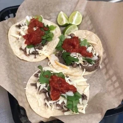 Elm Creek Beef Three tacos with shredded cheese, cilantro, and salsa, arranged on parchment paper with two lime wedges.
