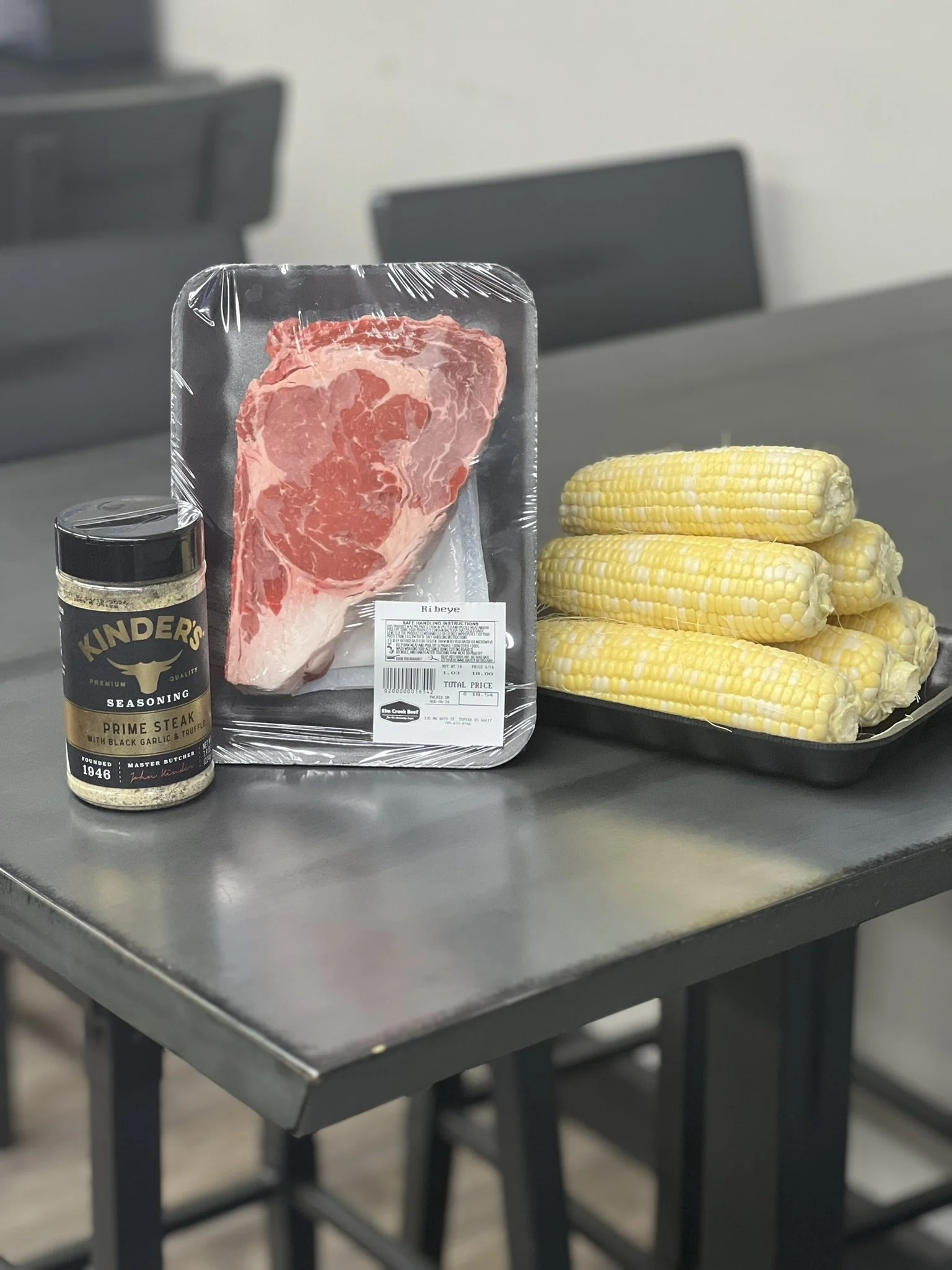 Elm Creek Beef Package of raw steak, a jar of Kinder's Prime Steak seasoning, and a tray of fresh corn on the cob on a black table.