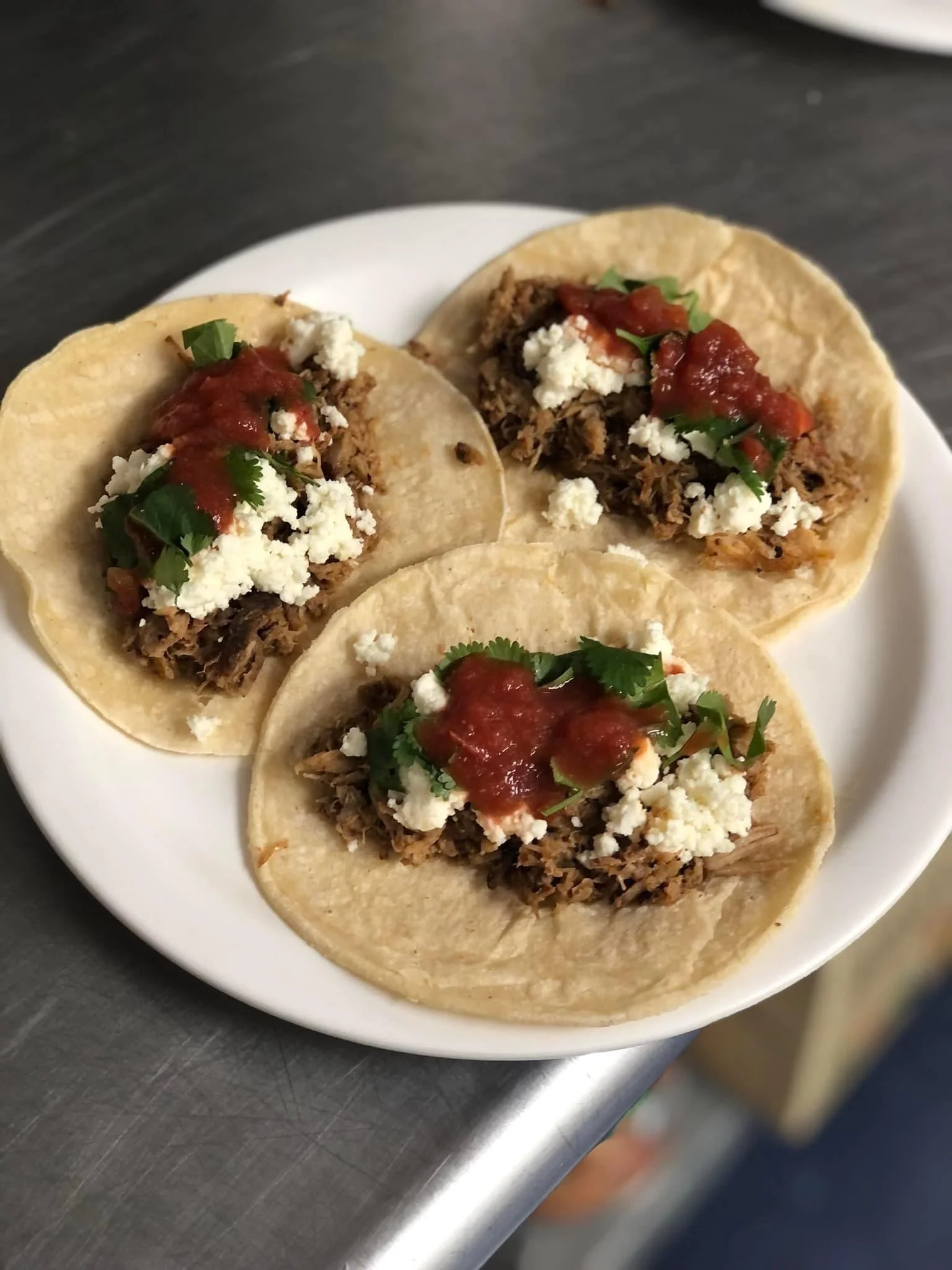 Elm Creek Beef Three tacos on a white plate with shredded meat, crumbled cheese, salsa, and cilantro, on a stainless steel surface.