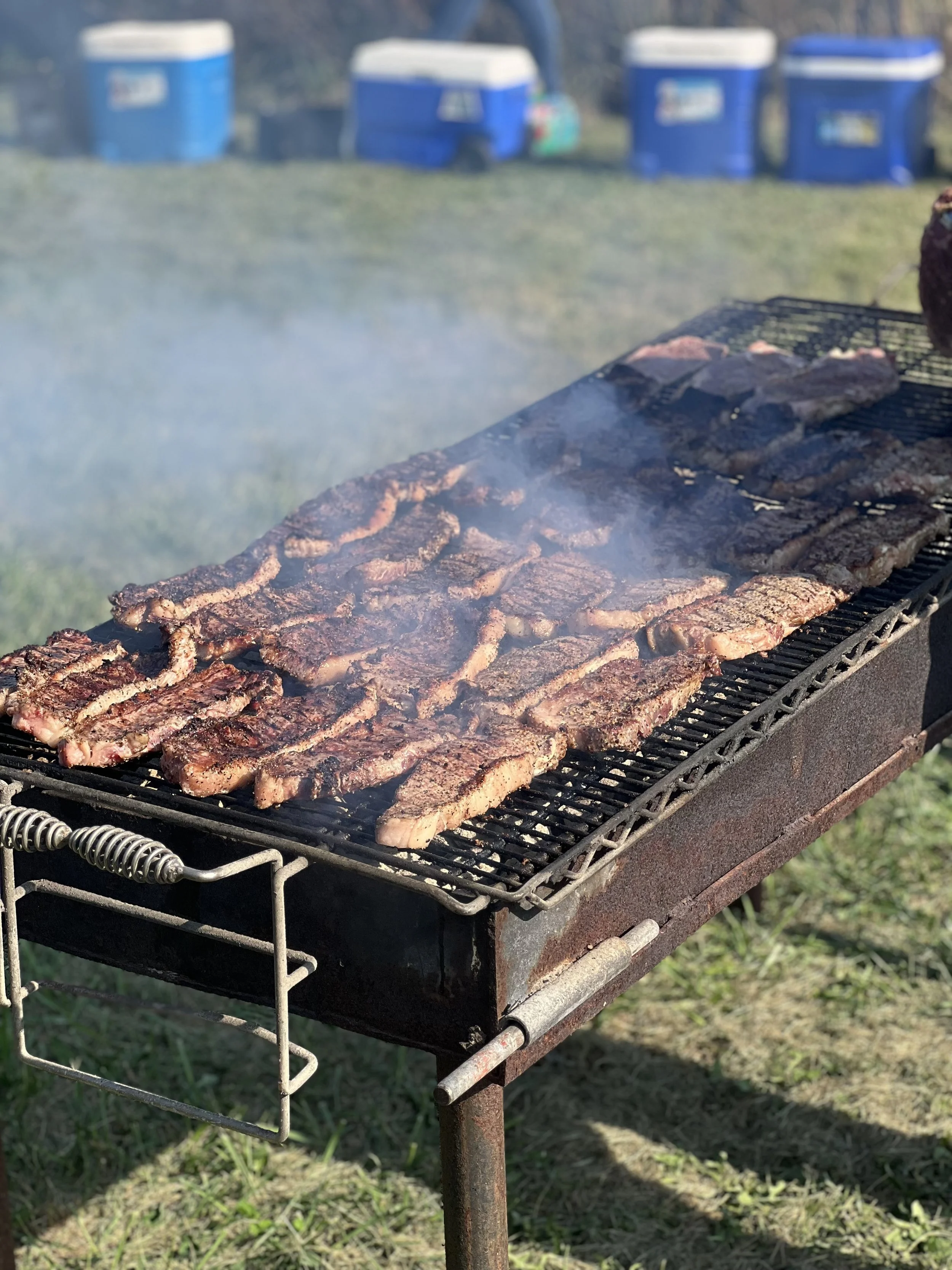 Barbecue grill with meat strips cooking on an outdoor grill, with smoke rising, and blue coolers in the background. Elm Creek Beef