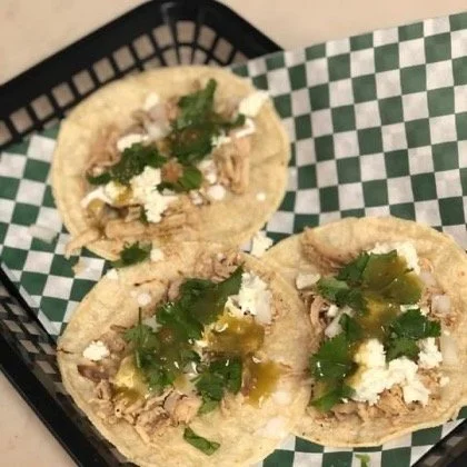 Elm Creek Beef Three tacos on a black tray with checkered green and white paper, filled with shredded chicken, chopped cilantro, onions, crumbled cheese, and topped with green salsa.