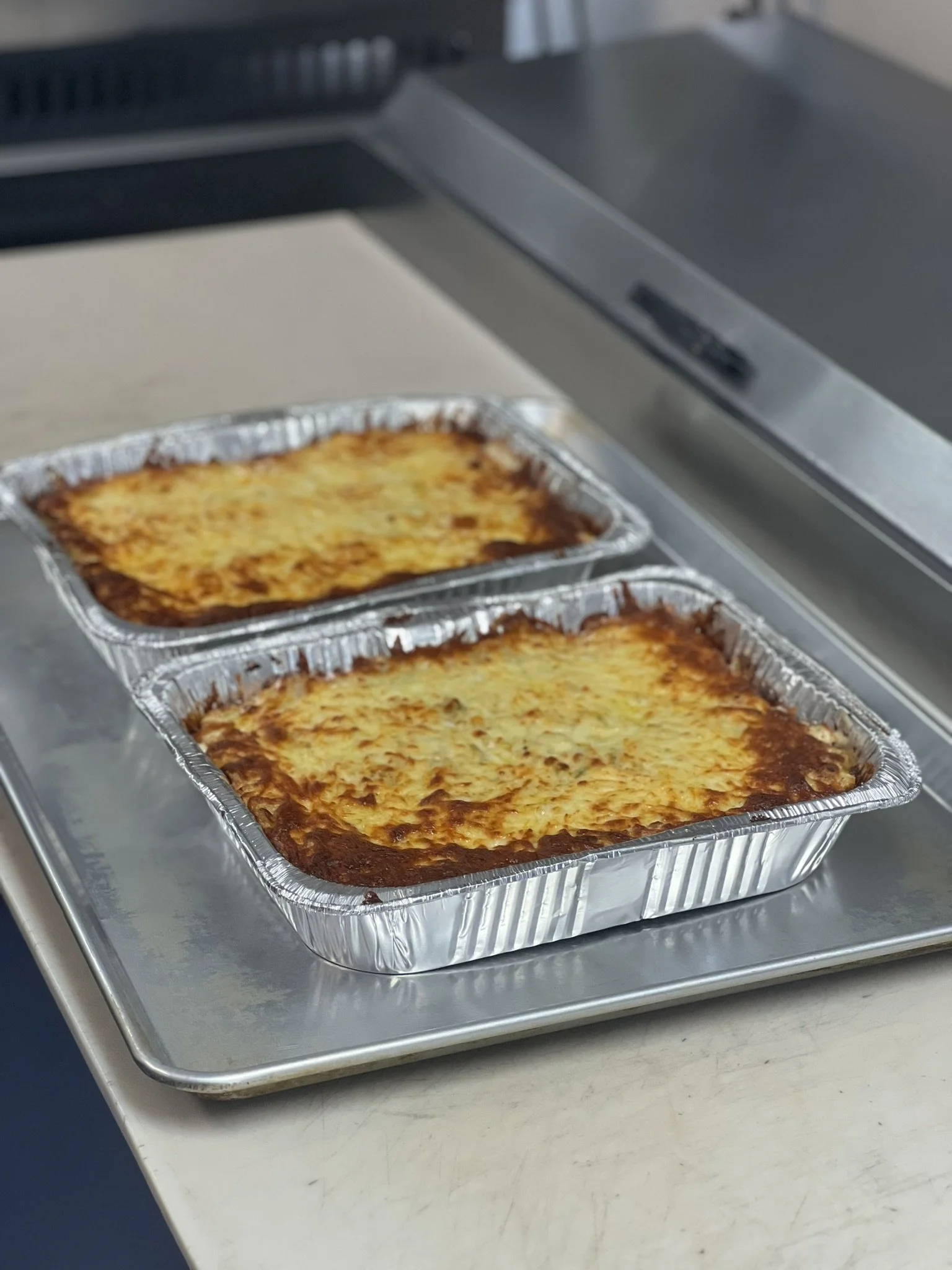 Elm Creek Beef Two takeout trays of baked lasagna on a metal tray in a kitchen.