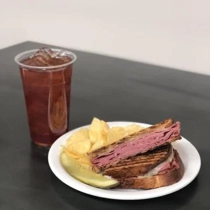 Elm Creek Beef A plate with a grilled steak sandwich, potato chips, and a pickle slice, alongside a glass of iced tea on a black table.