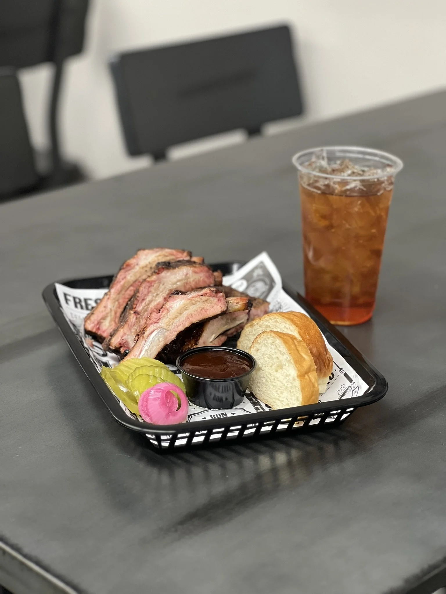 Elm Creek Beef A tray of brisket slices with barbecue sauce, bread rolls, pickles, and a pink onion slice, with a glass of iced tea in the background on a table.
