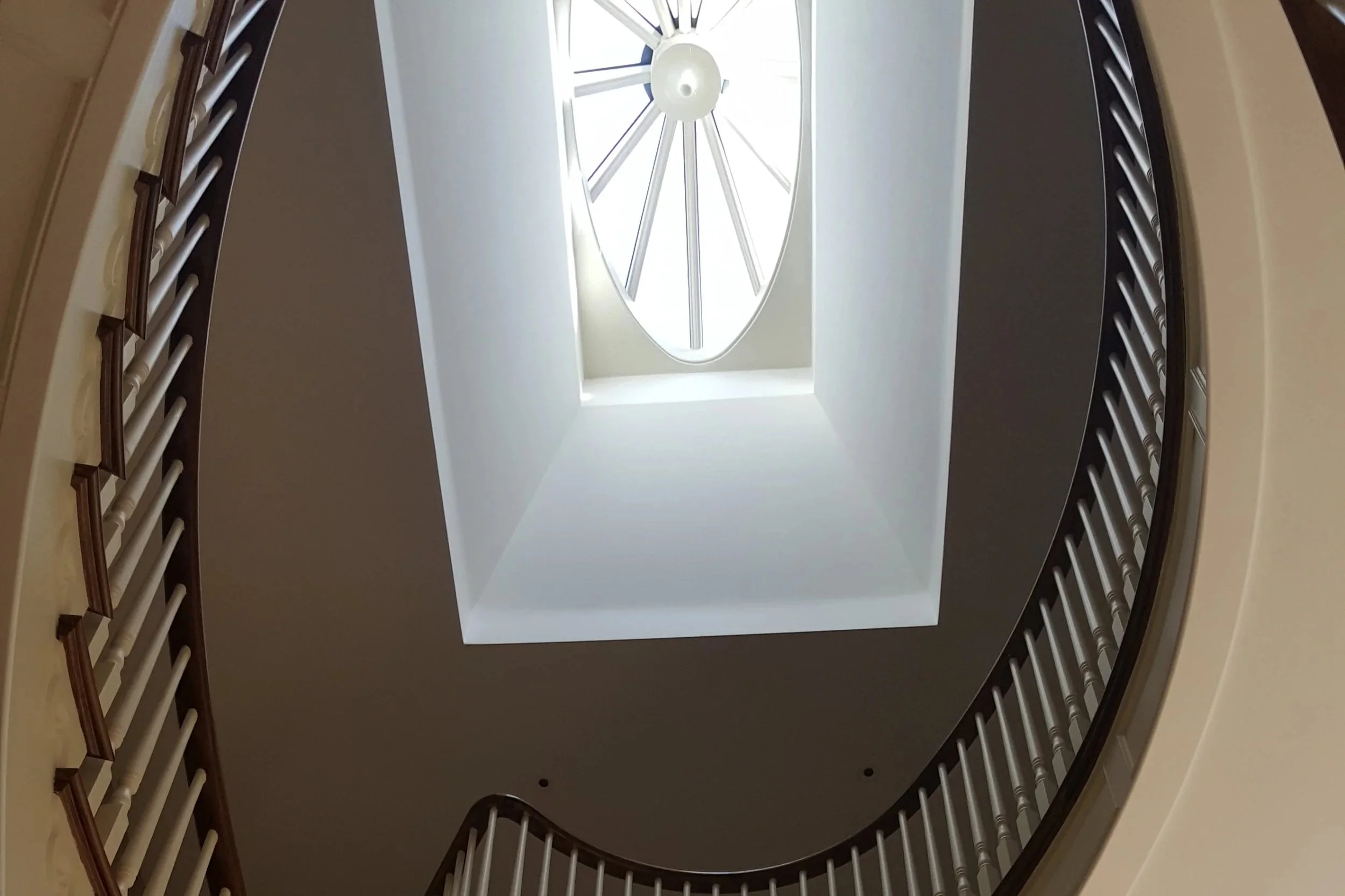 Looking up from the bottom of a spiral staircase inside a building, showing the ceiling with a large oval window and a modern light fixture, with railings along the staircase edges.