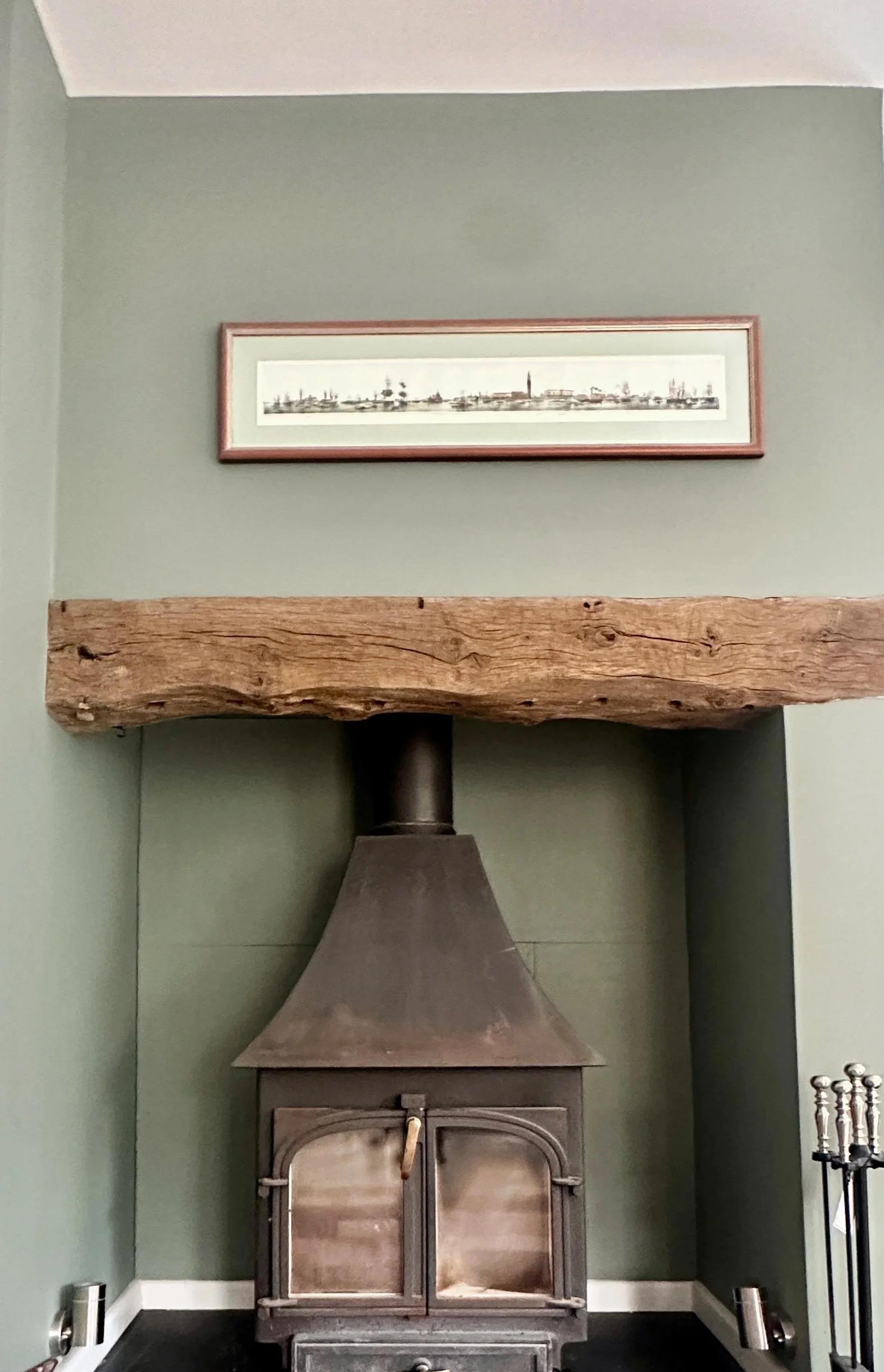 A room with green walls and a wood beam above a fireplace. Above the fireplace is a framed panoramic picture of a city skyline. A metallic sculpture and some candles are on the floor.