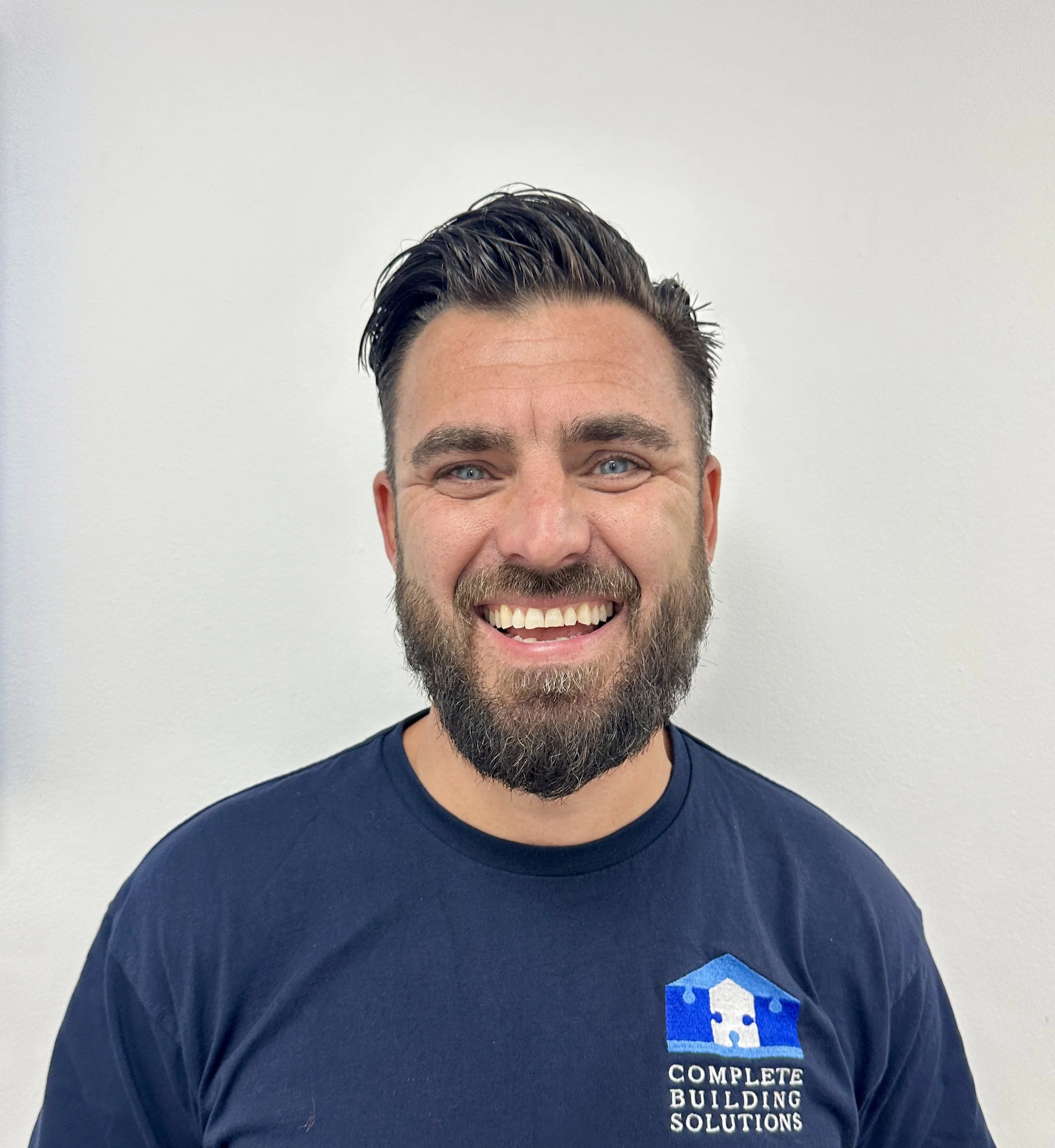 A smiling man with blue eyes, a beard, and styled dark hair, wearing a navy blue t-shirt with the logo and text 'Complete Building Solutions' on it, standing against a plain white wall.