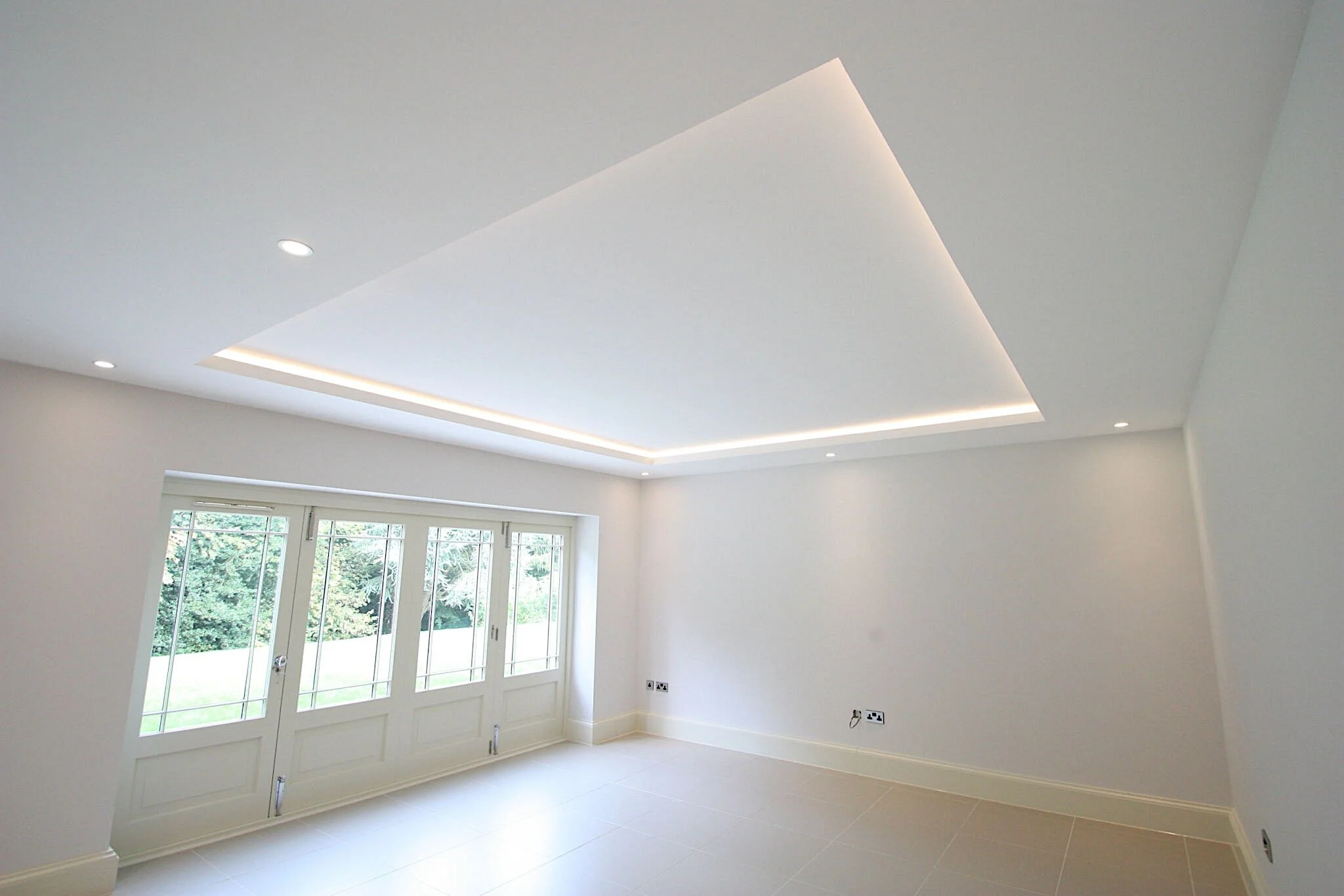 Empty white room with large windows, beige tile flooring, white walls, and a ceiling with recessed lights and indirect lighting.