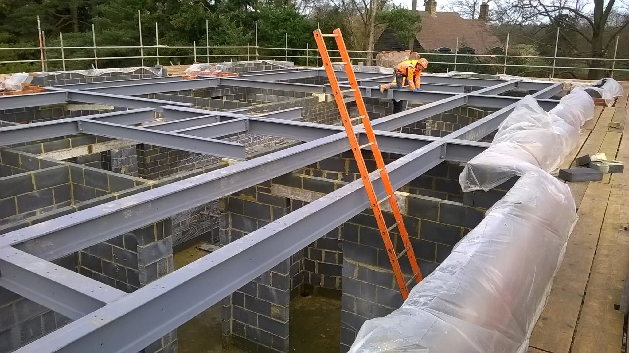 Construction workers install a steel framework on top of brick walls for a building. An orange ladder leans against the framework, and plastic sheeting covers parts of the structure. The area is surrounded by safety railings, and surrounding trees an