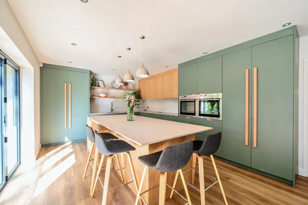 Modern kitchen with green cabinets, wooden accents, and a large island with four bar stools. Light wood flooring and natural light from a sliding glass door.