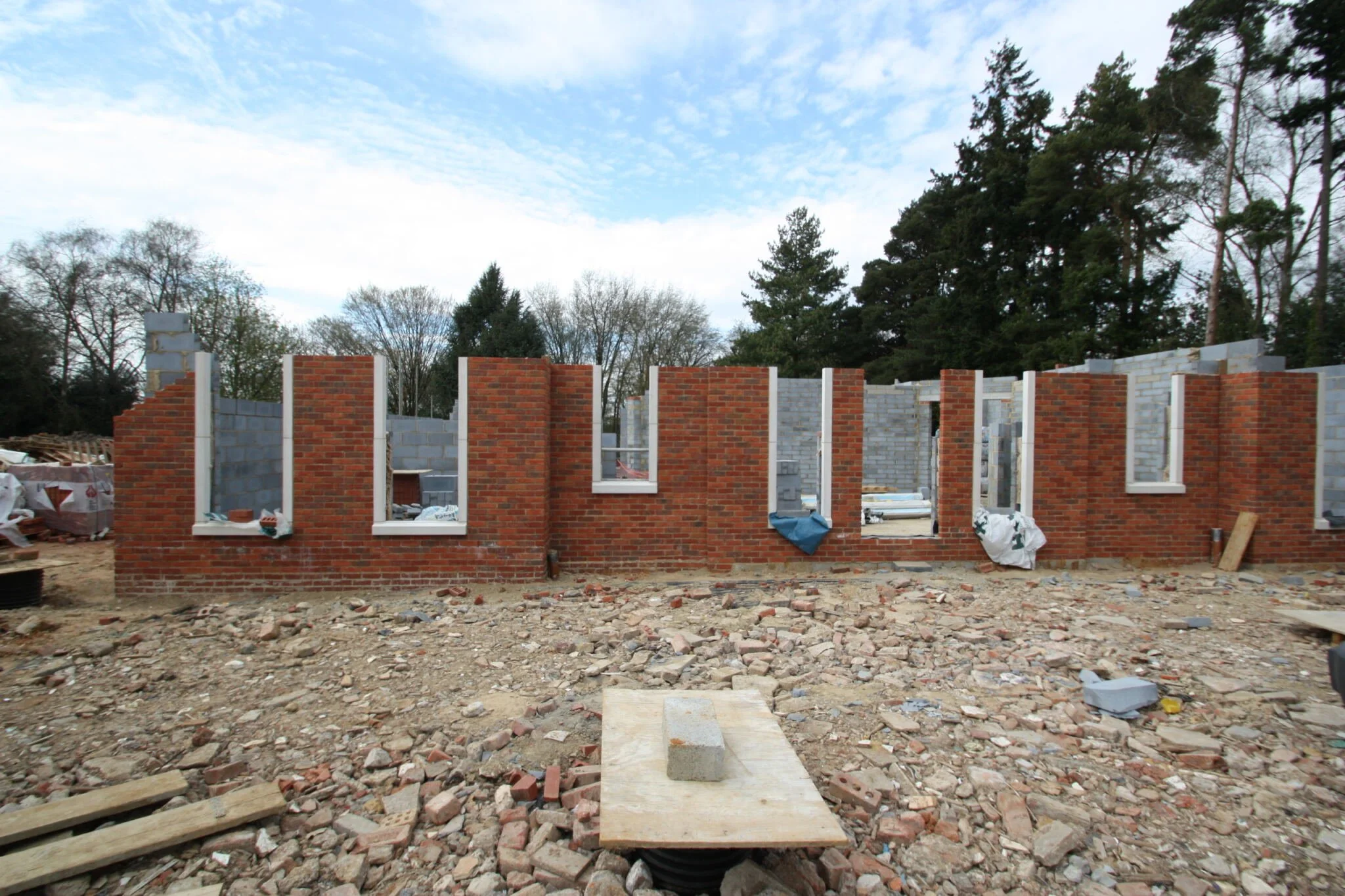 Under construction brick building with open window spaces, construction debris on ground, and trees in background.
