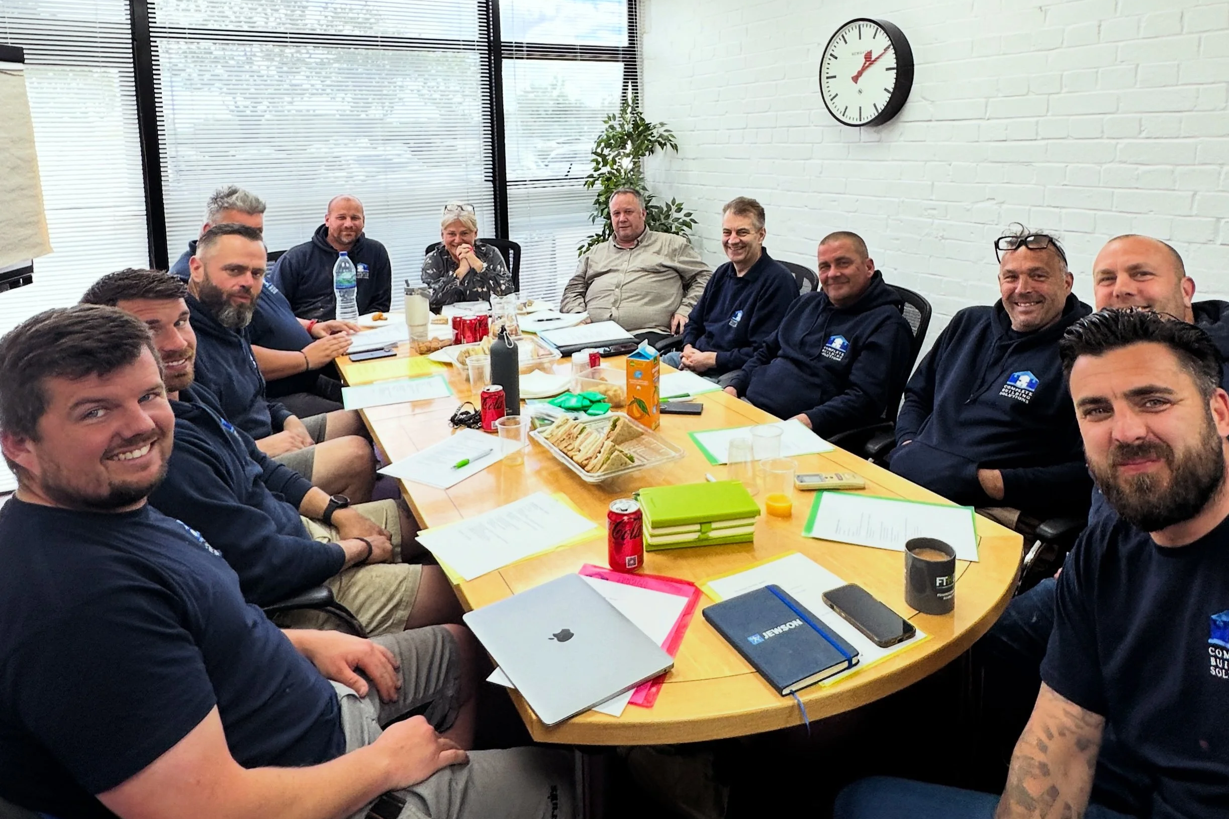 A group of people gathered around a conference table in a woking office, smiling at the camera, with snacks and drinks on the table and a large clock on the white brick wall.