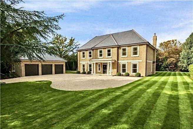 A large two-story beige house with a gray roof, surrounded by a manicured lawn with stripes, and a three-car garage on the left. Trees are in the background, and the sky is partly cloudy.