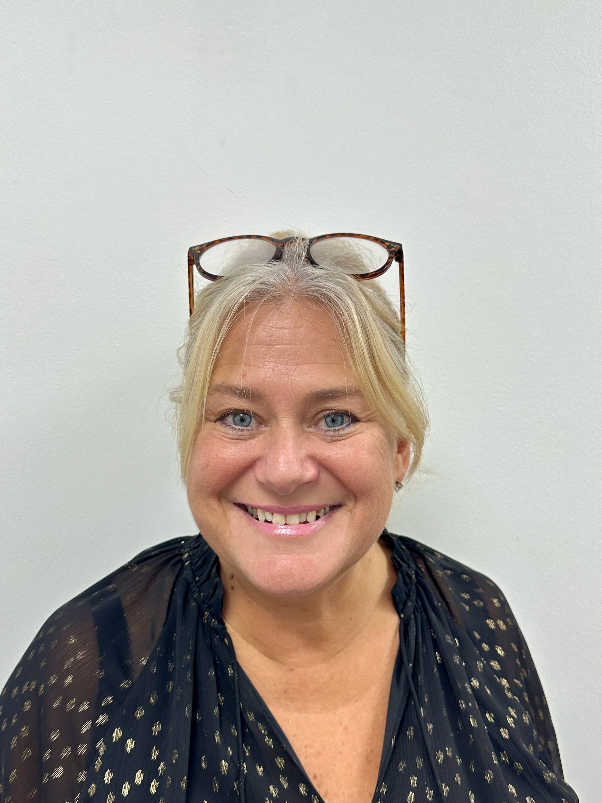 Becky with short blonde hair, blue eyes, and light skin, smiling and wearing pink glasses on her head, sitting in an office with blinds and stacks of files and papers behind her.