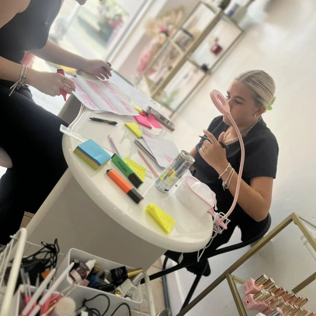 A woman sitting at a cluttered desk examining her makeup. The desk has various colored sticky notes, markers, and documents. The background includes shelves with beauty products and a window with a view outside.