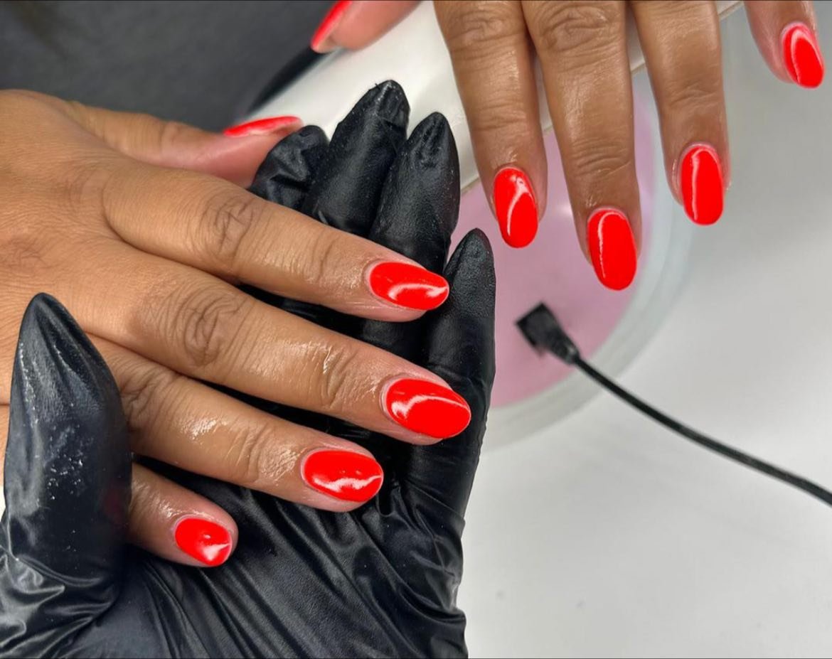 A person getting red gel nail polish applied to their fingernails by a professional wearing black gloves.