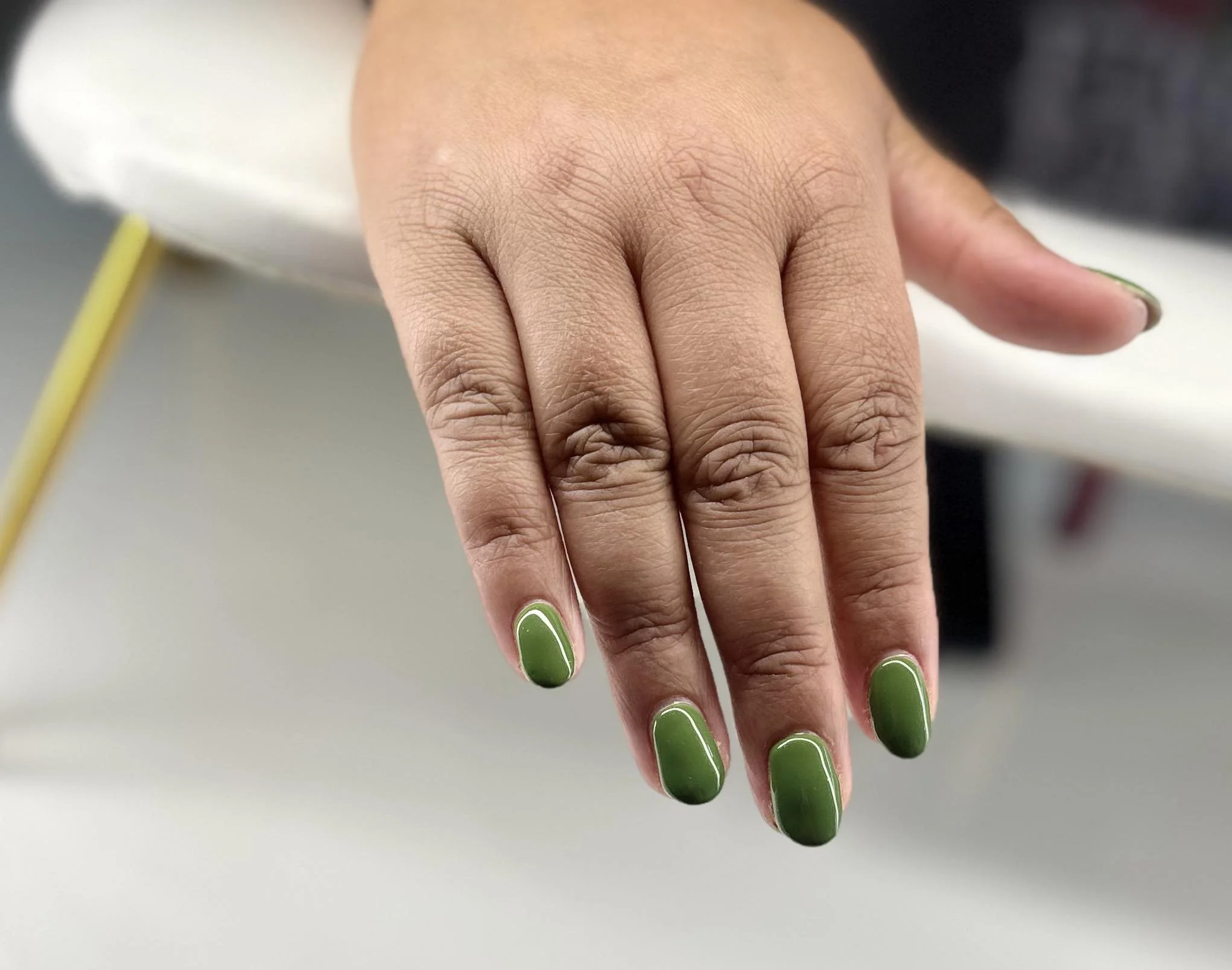 Close-up of a hand displaying nails painted in dark green with a glossy finish. The hand rests on a surface with a blurred background.