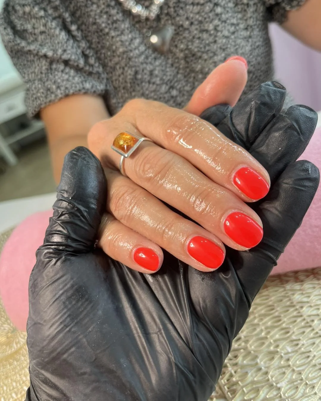 A person receiving a manicure with bright red nail polish, wearing a silver ring with amber, while their hand is held by a person in a black glove.