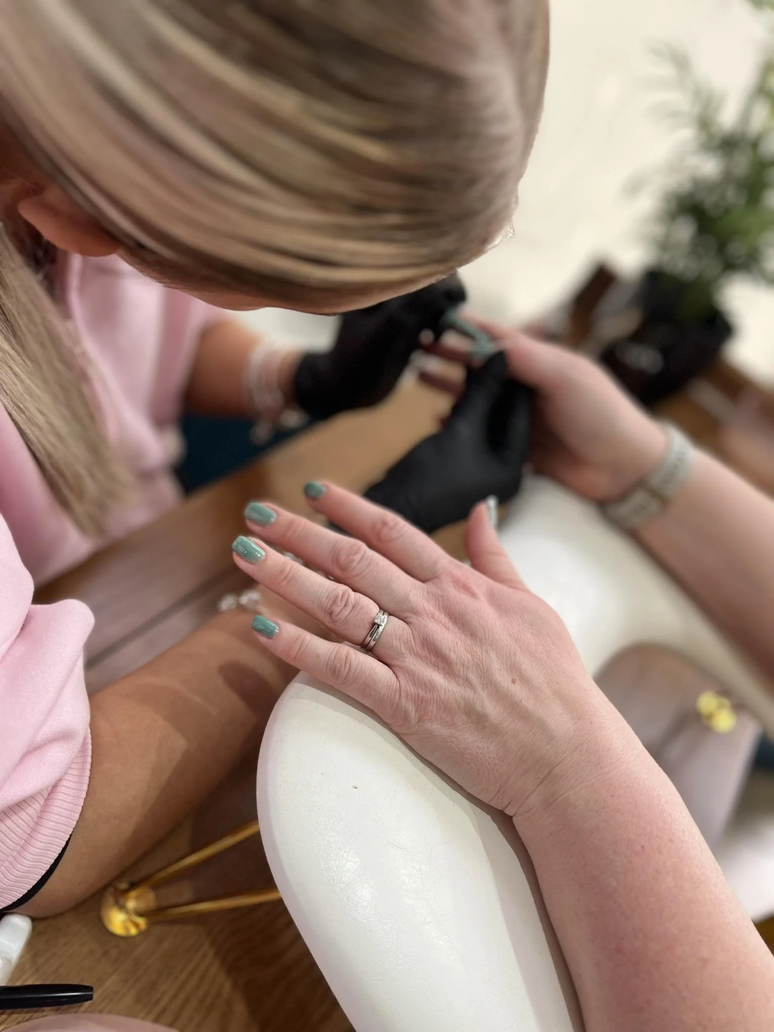 A person receiving a manicure, with nails being painted while their hand is resting on a white surface. The manicurist is wearing black gloves and is focused on the task.