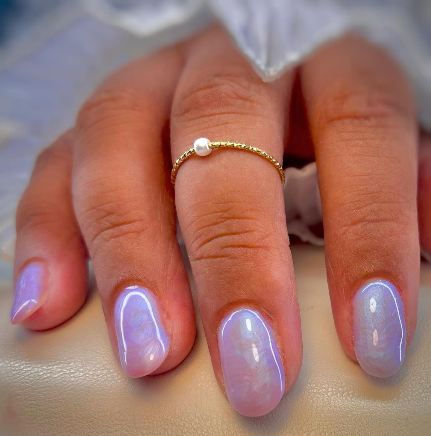 A hand with manicured nails painted in a pastel purple color, wearing a delicate gold ring with a single pearl, resting on a beige surface.