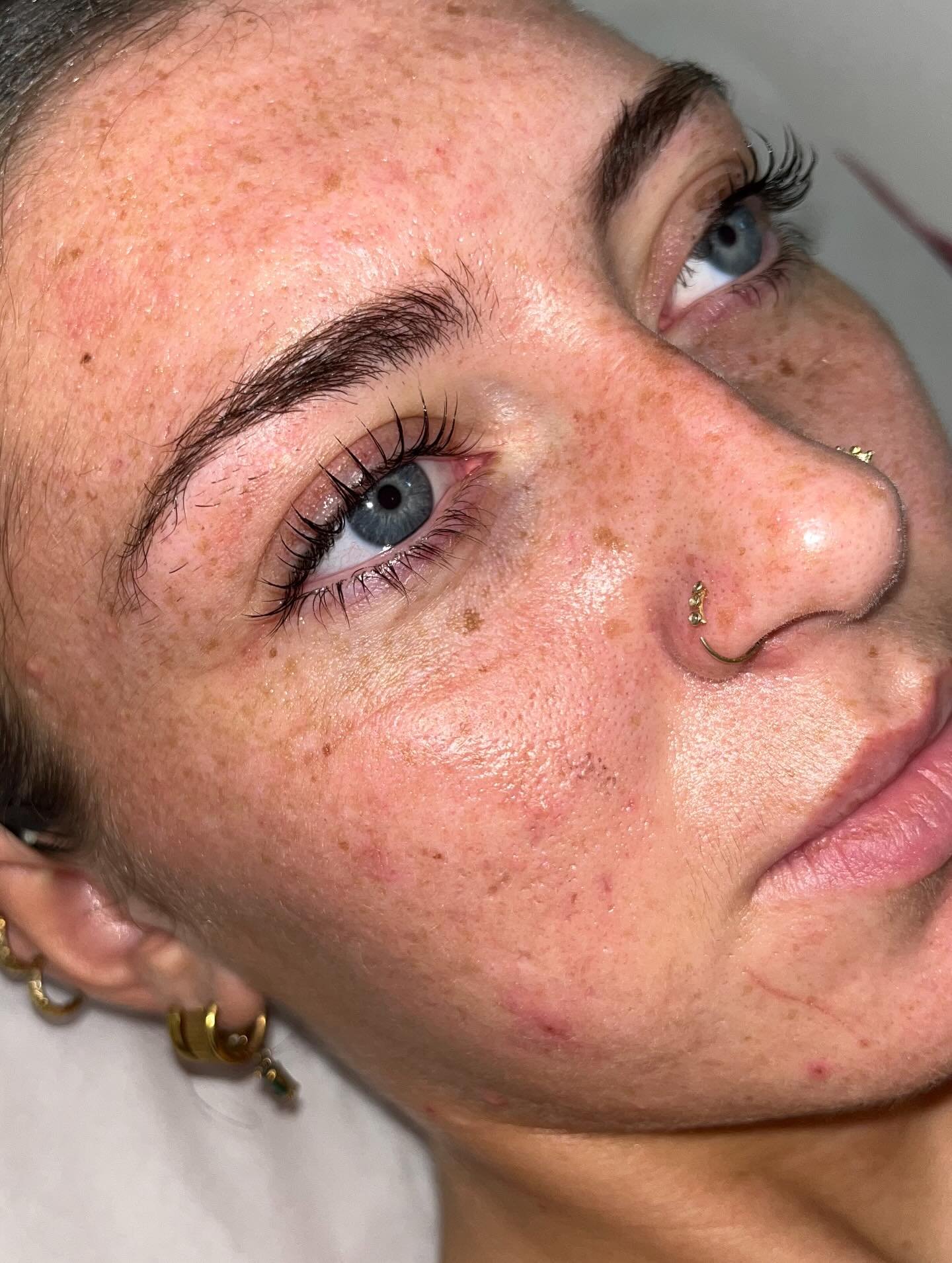 Close-up of a woman's face showing clear skin with freckles, blue eyes, and gold jewelry including a nose ring and earrings.