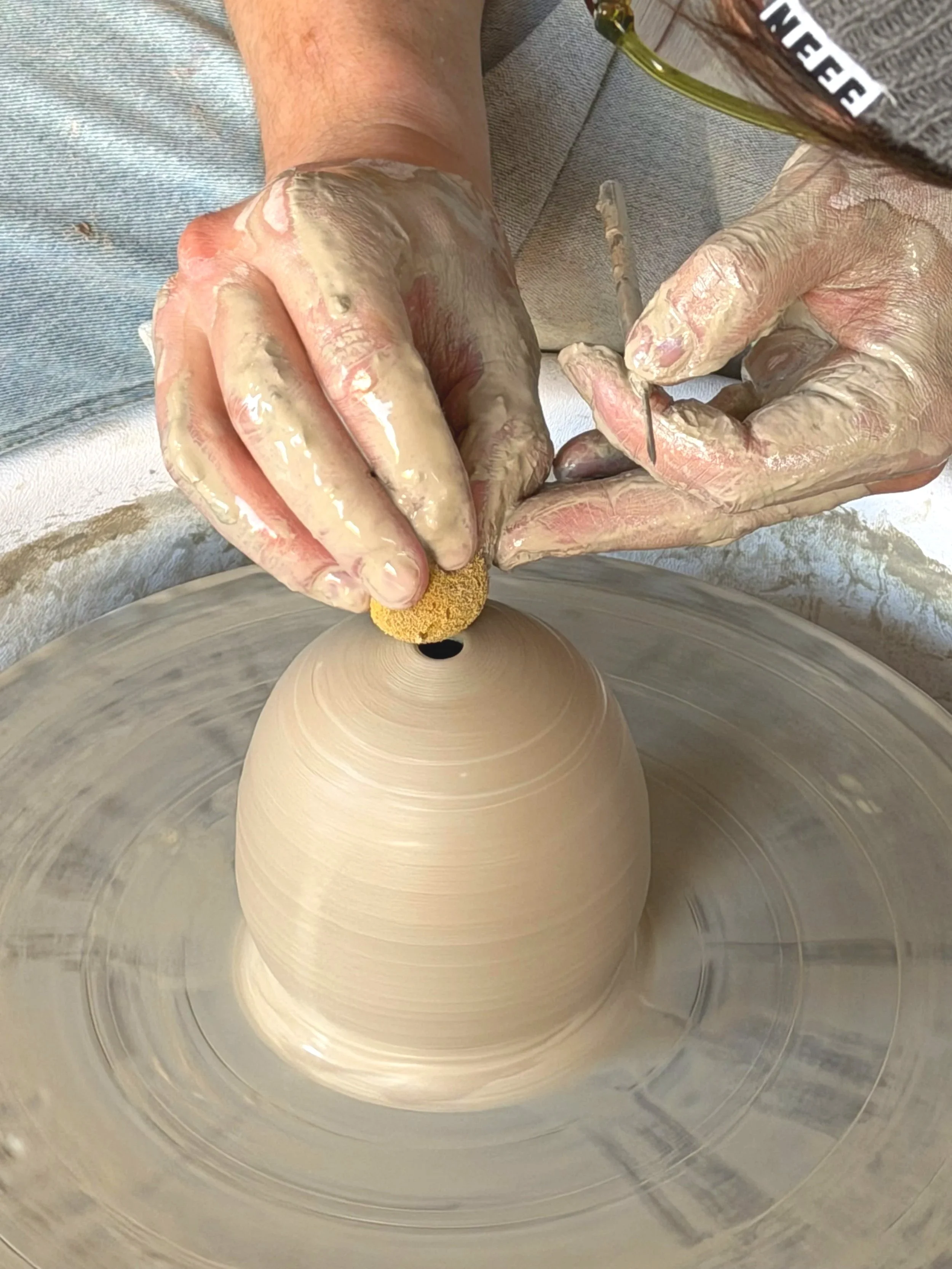 Person shaping a clay vase on a pottery wheel with hands covered in wet clay.