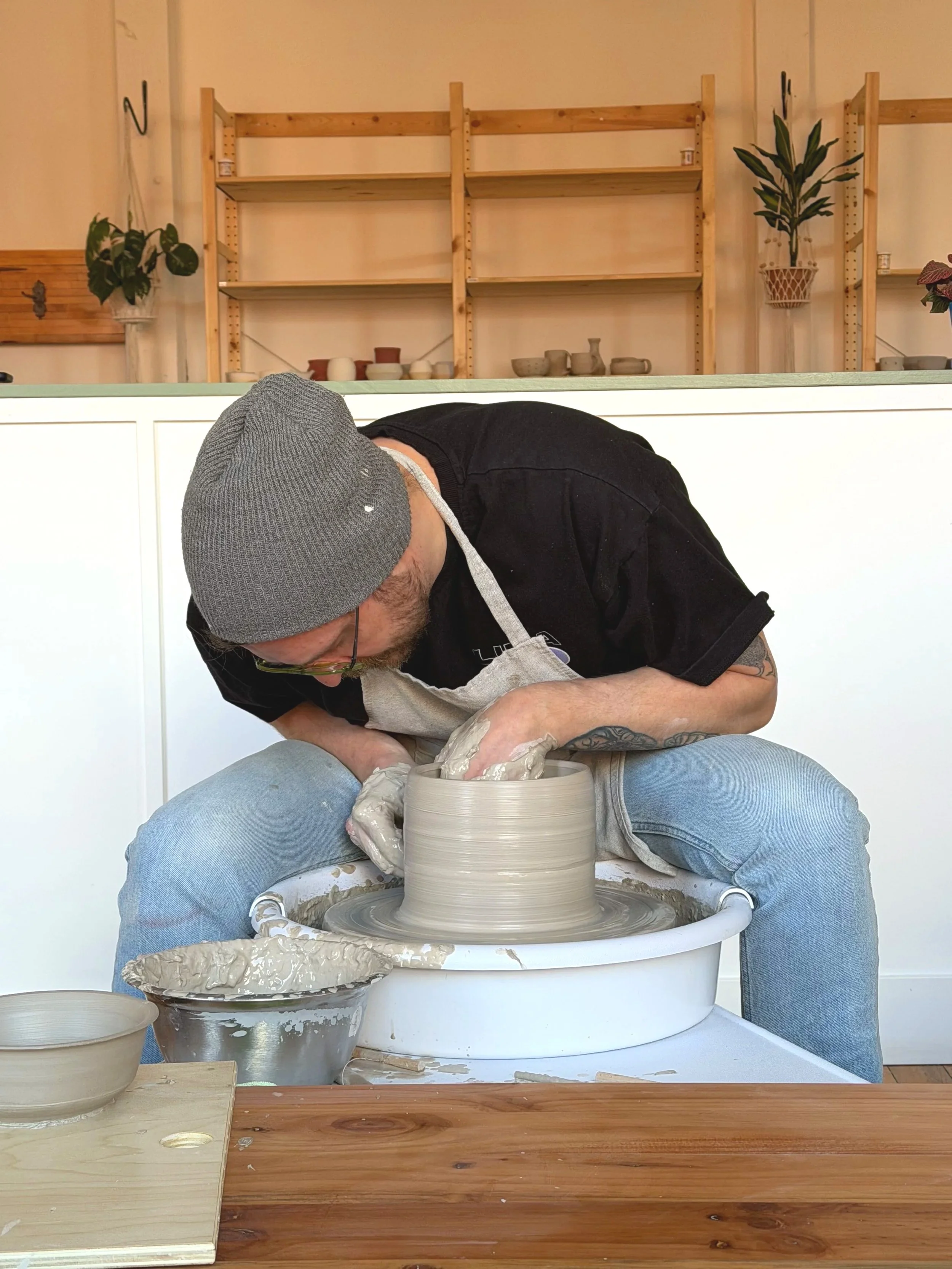Man wearing a gray beanie and glasses, dressed in a black shirt and jeans, is pottery making on a pottery wheel in a pottery studio with wooden shelves and potted plants in the background.