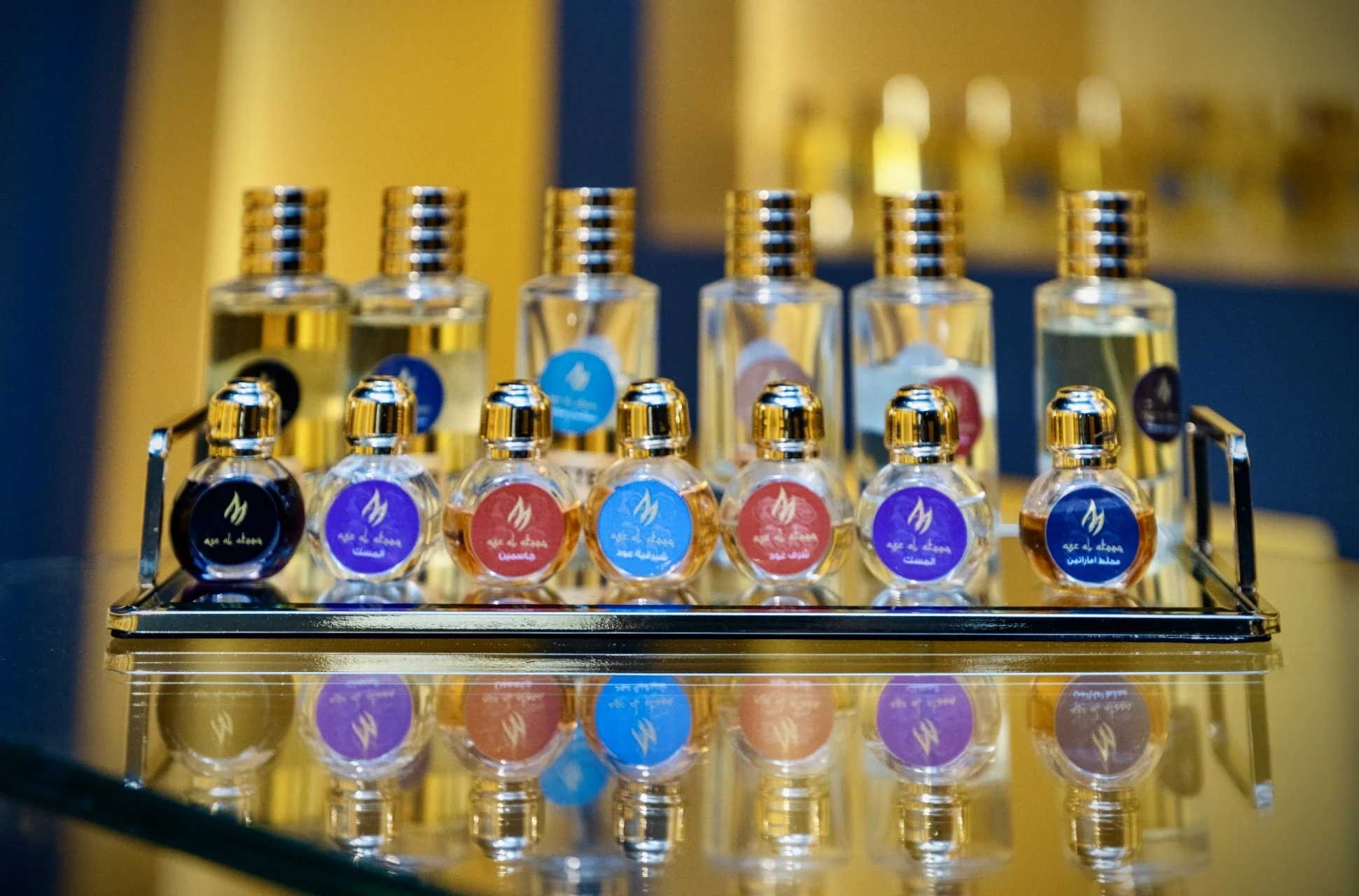 A display of small Oud bottles with colorful labels on a reflective surface. The bottles have gold caps and are arranged on a metal tray.