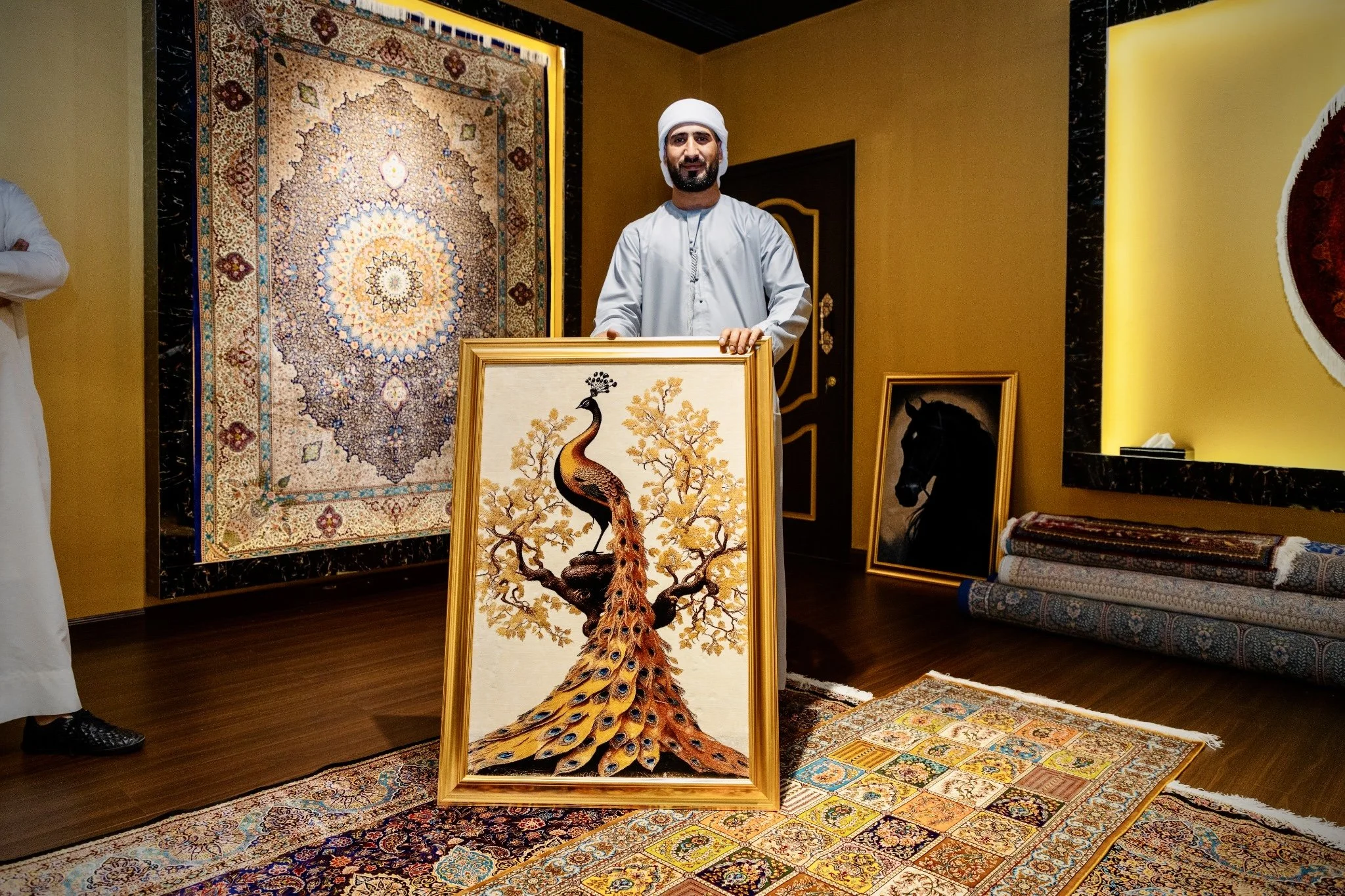A man in traditional Middle Eastern attire holds a framed artwork of a peacock with long tail feathers, inside an art gallery with various handcrafted Carpets and artwork, including a large, elaborate Carpet hanging on the wall behind him.
