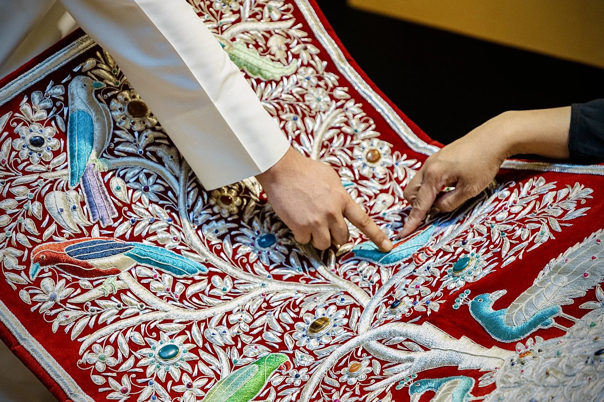 Close-up of embroidery on red fabric, featuring colorful peacocks, flowers, and leaves with intricate detailing and bead accents.