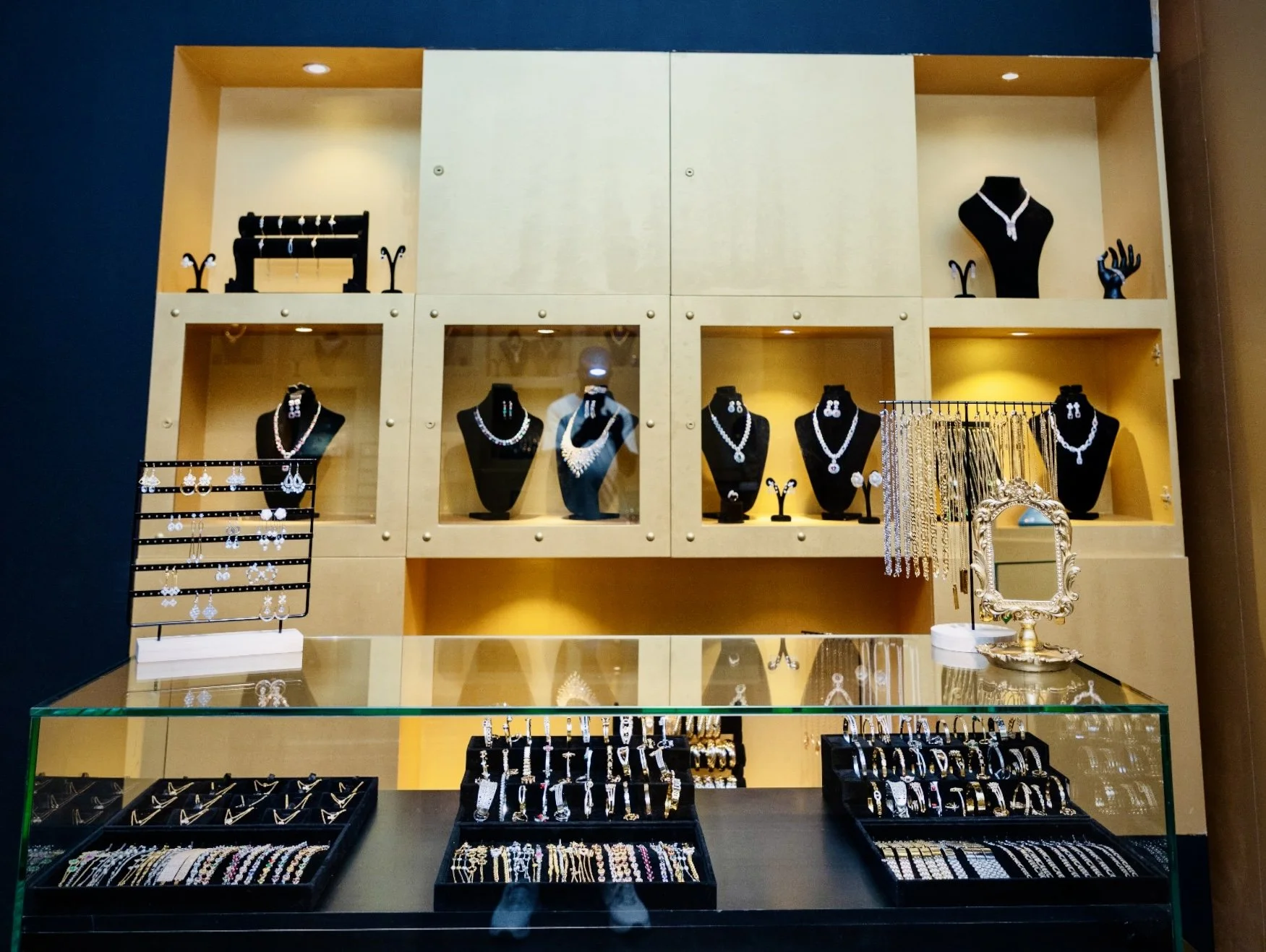 Jewelry display with necklaces, earrings, rings, and bracelets in a glass case, with additional jewelry on black busts and stand, and a decorative mirror on the right.
