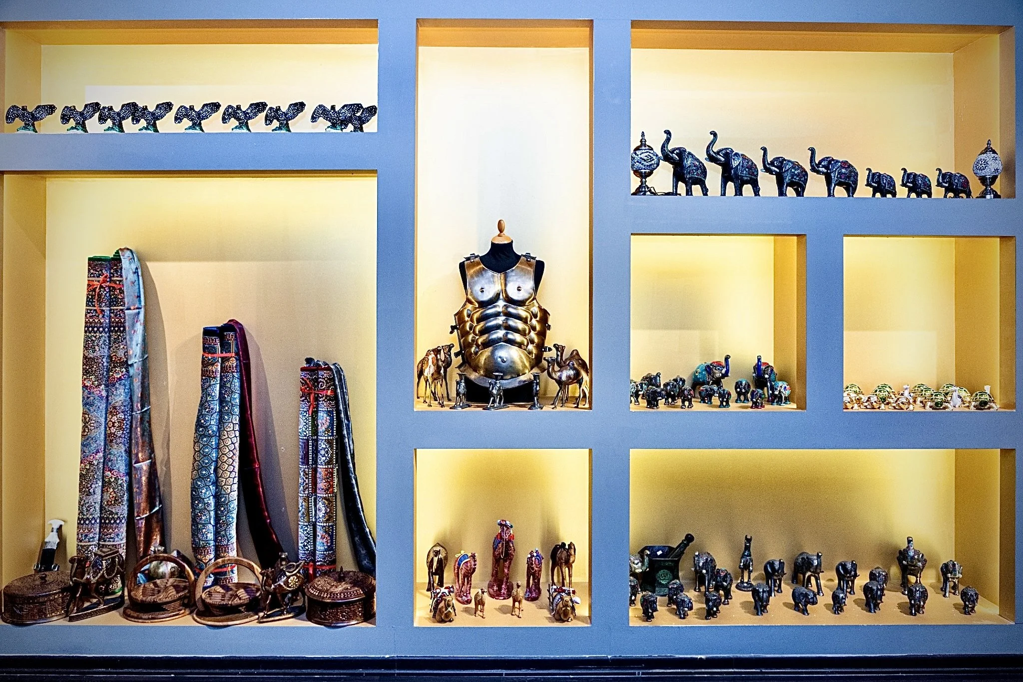 Wooden shelf display with small colorful elephant figurines, decorative vases, and striped textiles, with yellow and blue background lighting.